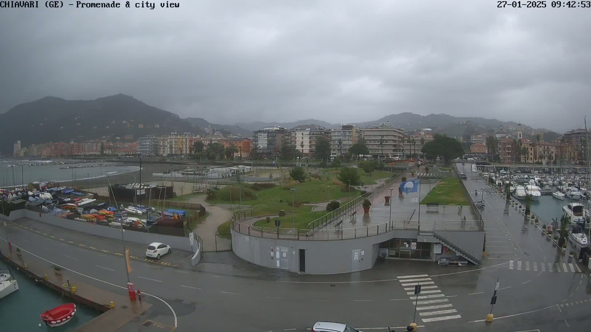 A wet, overcast day shows a harbor with many boats, a promenade, and a city backdrop of multi-story buildings nestled at the base of hills.