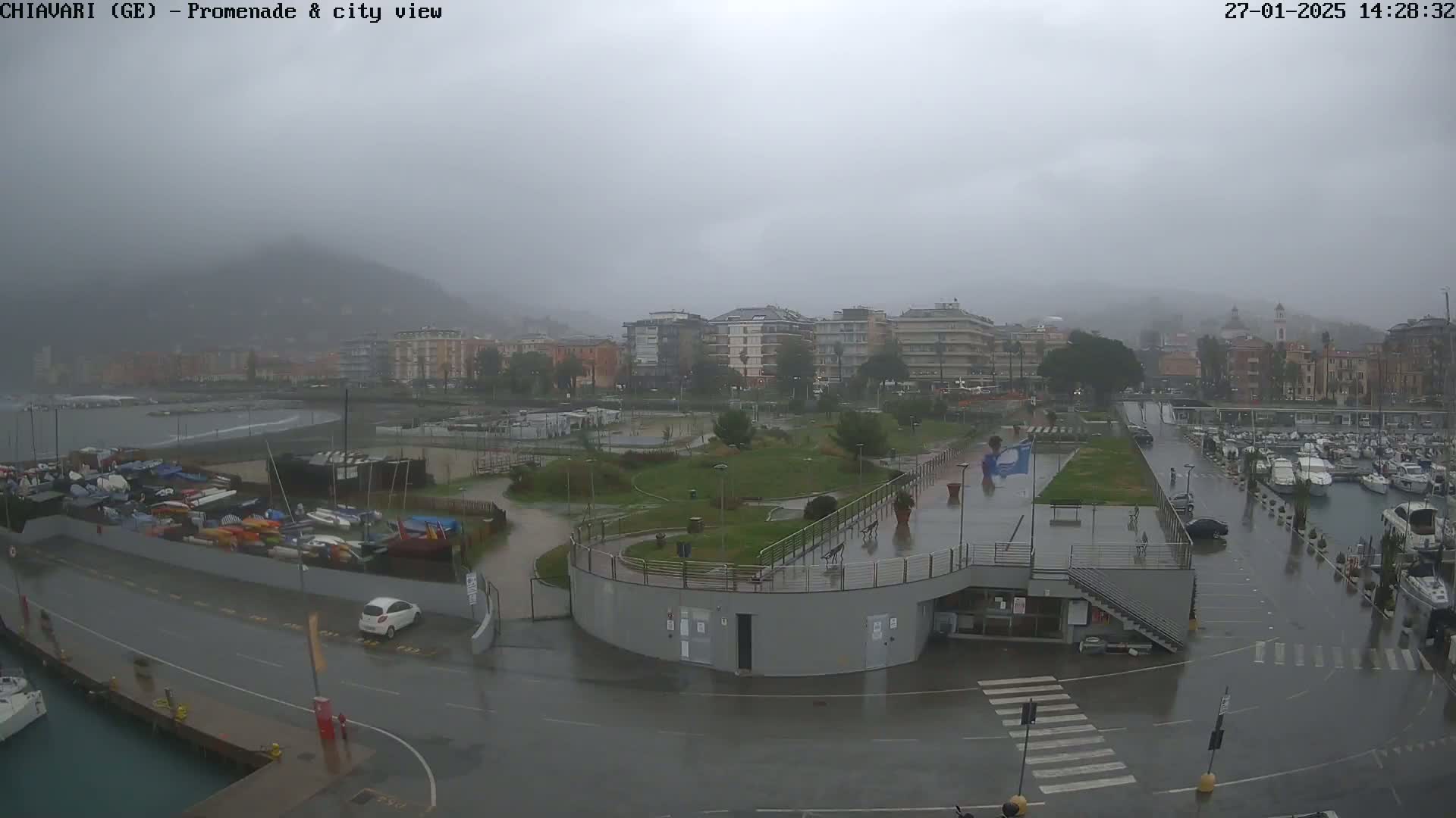 A rain-soaked waterfront promenade and marina, with a city and hills in the background under an overcast sky.
