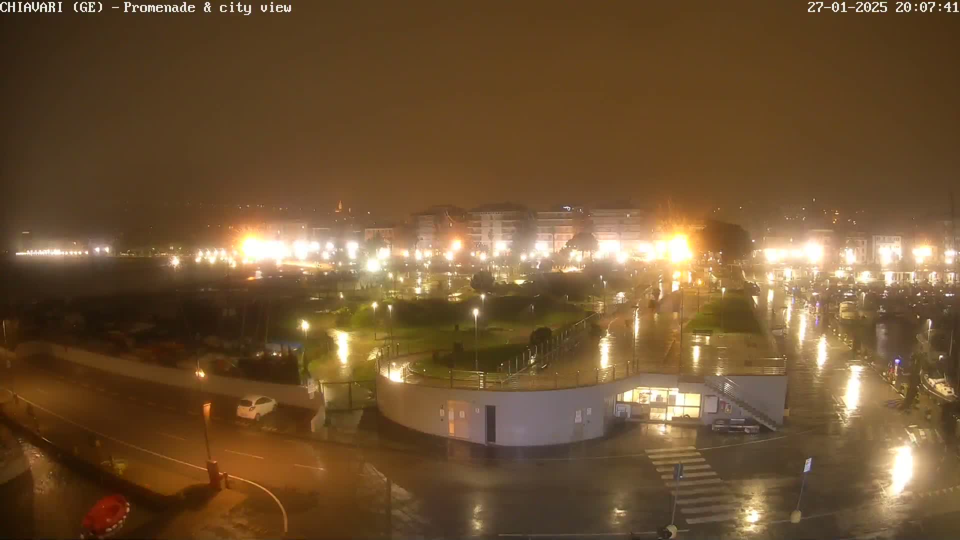 A rain-slicked waterfront promenade and city at night, illuminated by streetlights reflecting in the wet surfaces.