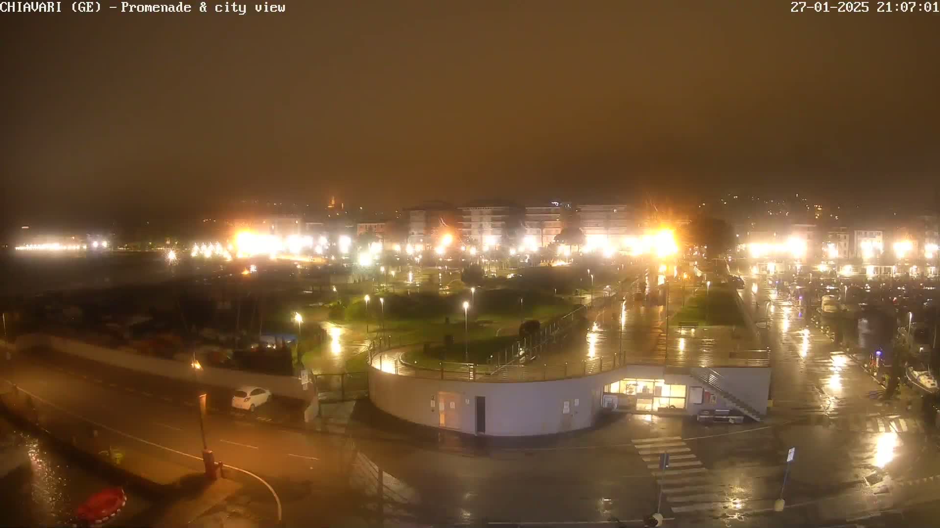 A rain-slicked, nighttime view of a city waterfront promenade and harbor, illuminated by streetlights.
