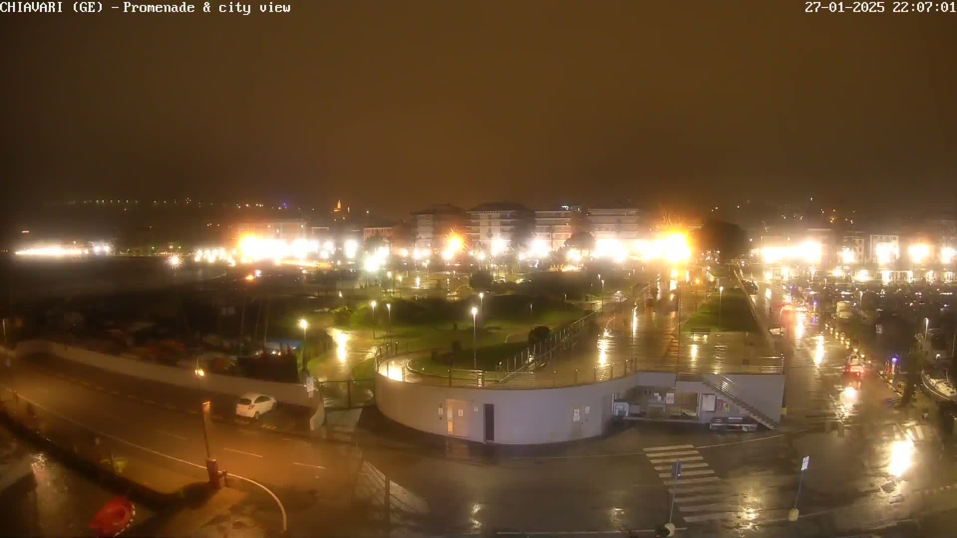 A nighttime view of a rain-slicked city promenade and harbor, illuminated by streetlights and building lights.