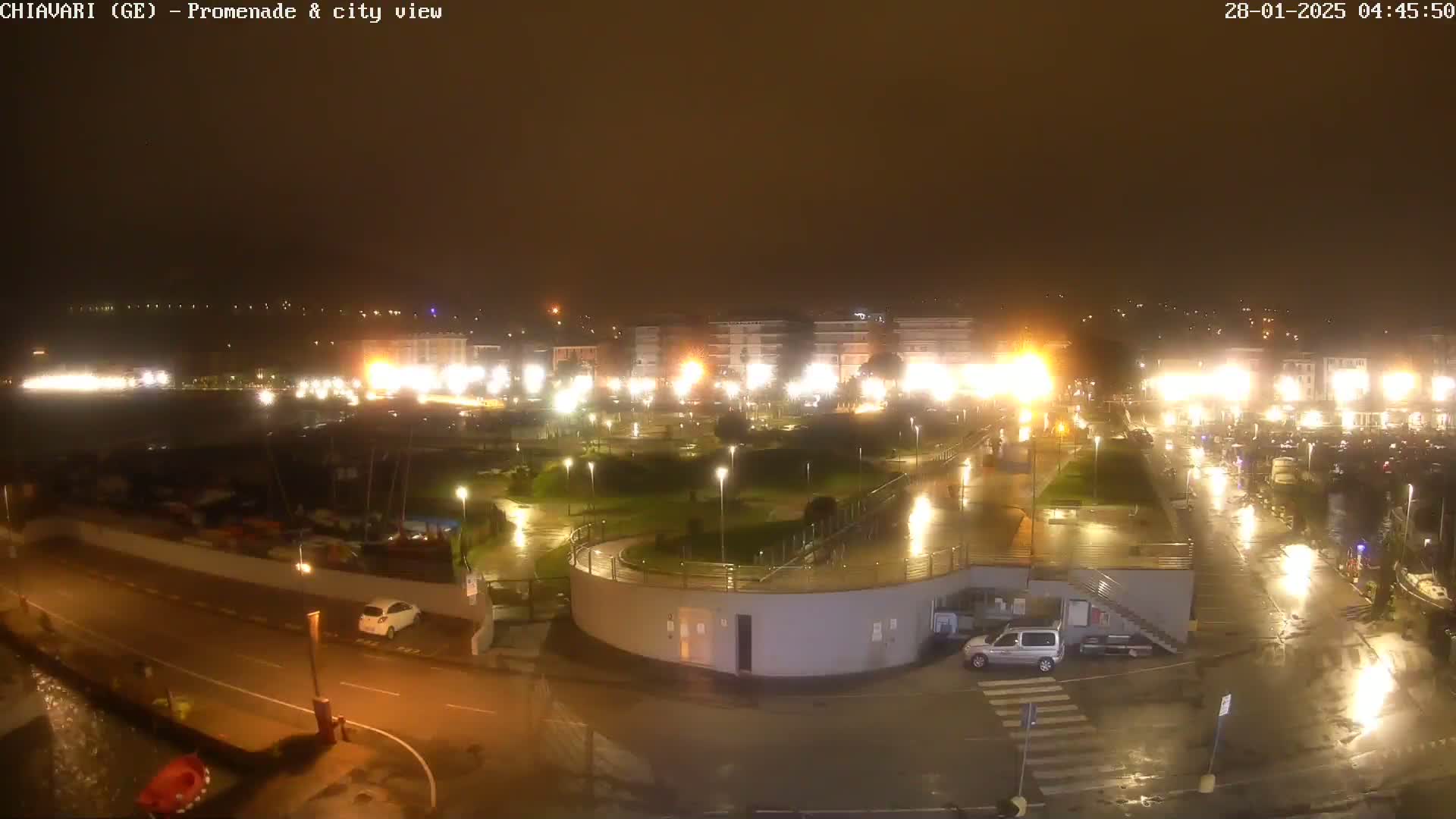 A nighttime view of a rain-slicked city promenade and harbor, illuminated by streetlights and building lights.