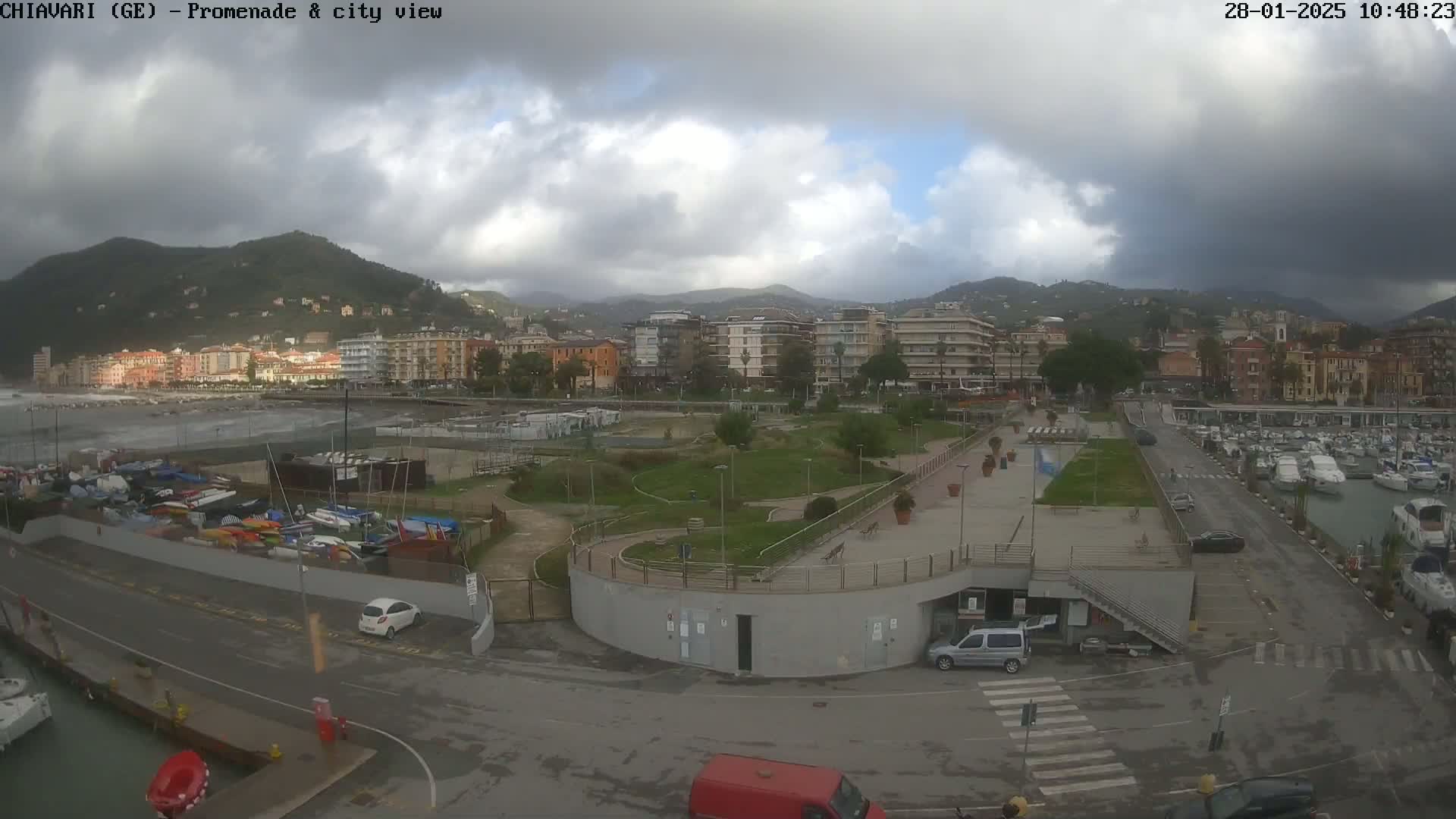 A coastal city scene under a cloudy sky shows a promenade, a marina with boats, and buildings nestled against hills in the background.