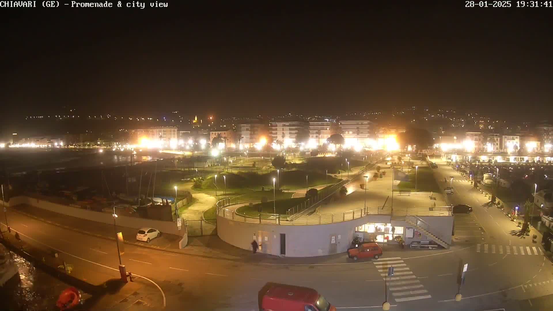 A nighttime aerial view of a coastal town's promenade and city, illuminated by streetlights, showing a park, roads, and parked cars.