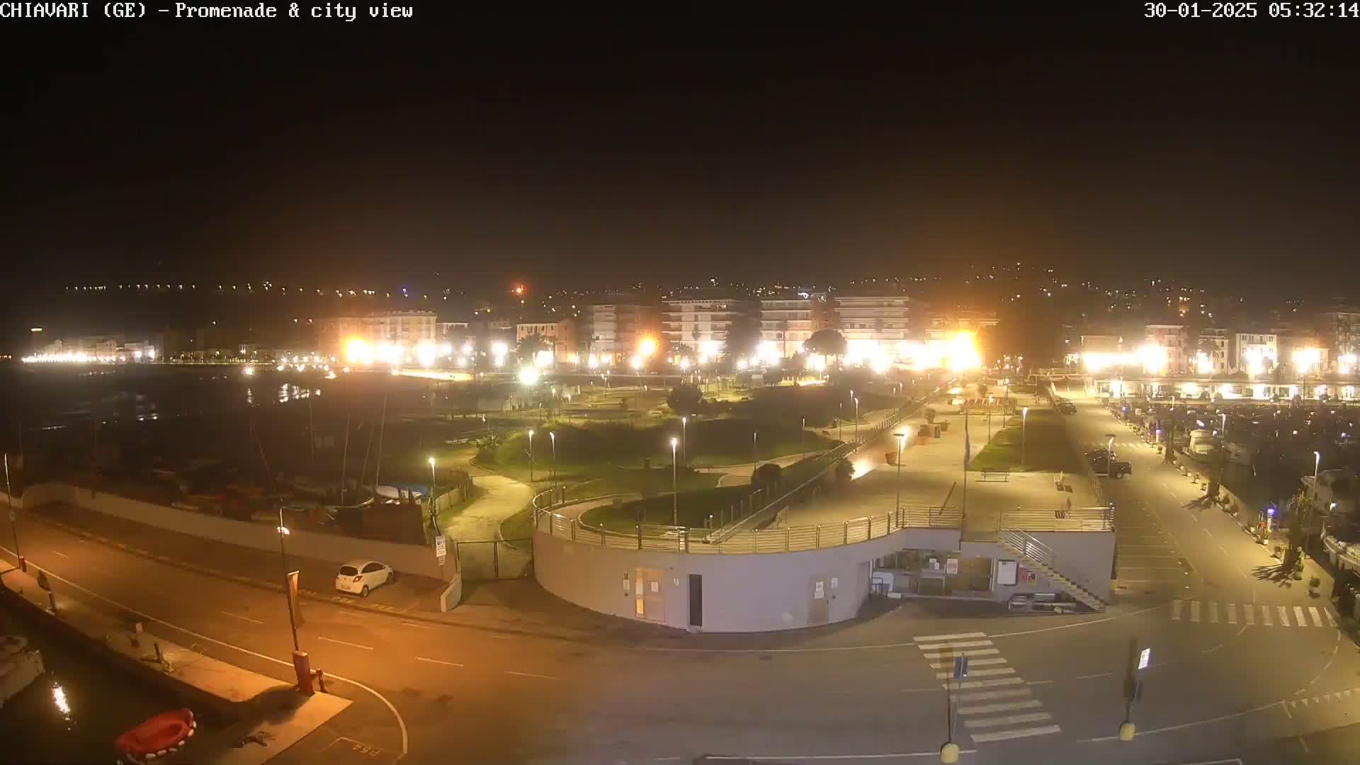 A nighttime aerial view of a city's illuminated waterfront promenade and harbor, featuring a park and parking areas.