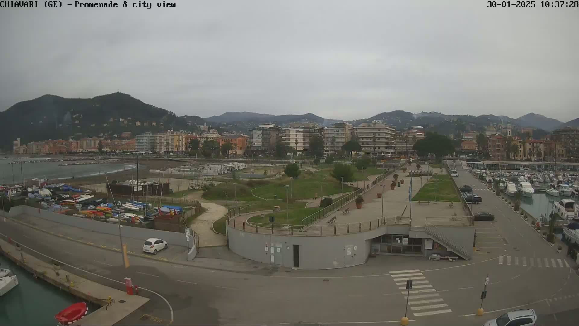 A coastal city with a harbor full of boats is viewed from above on an overcast day, showing a promenade and park alongside a road.