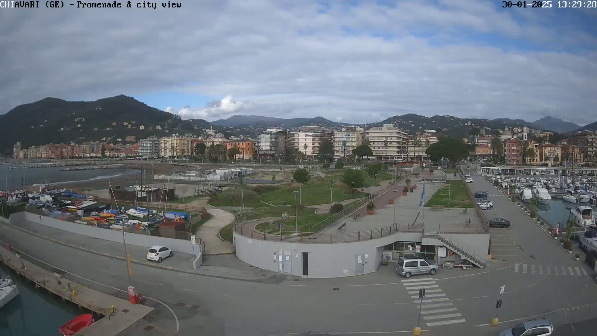 A coastal city with a marina and promenade is visible under a partly cloudy sky, with mountains in the background.