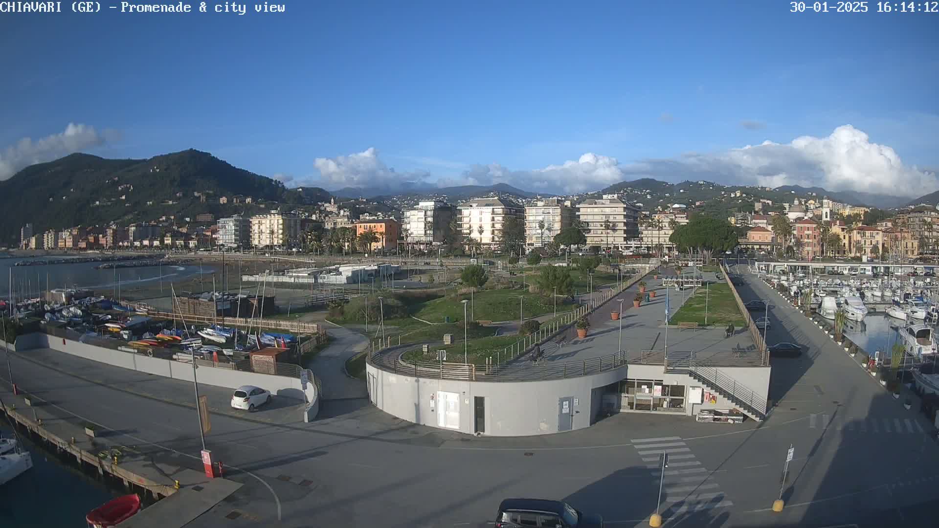 A coastal city with buildings, a marina filled with boats, and a paved promenade with a park under a partly cloudy sky.