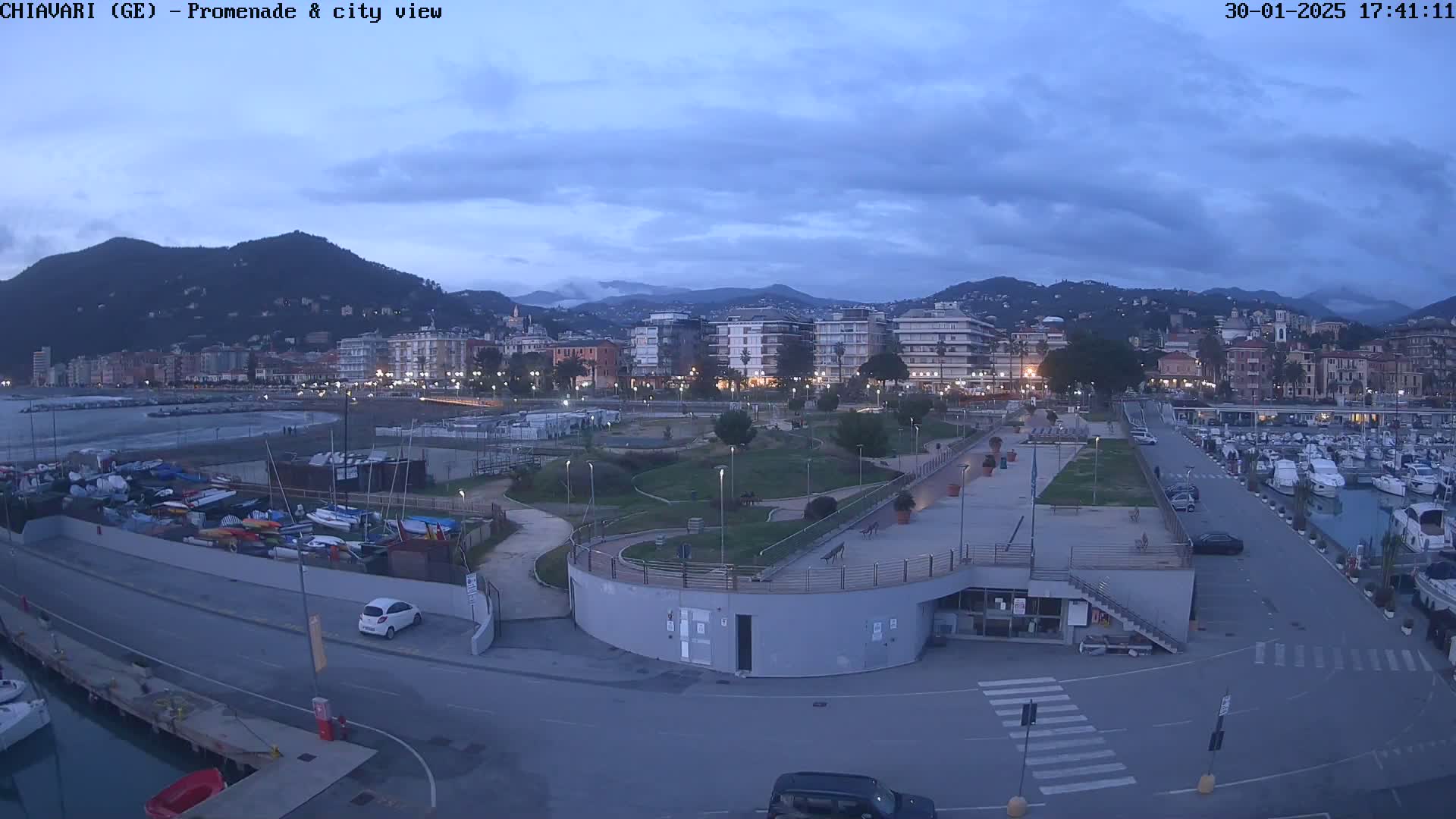 An aerial view of a harbor city at dusk, showing a marina filled with boats, a promenade with a park, and buildings nestled against a hillside under a cloudy sky.