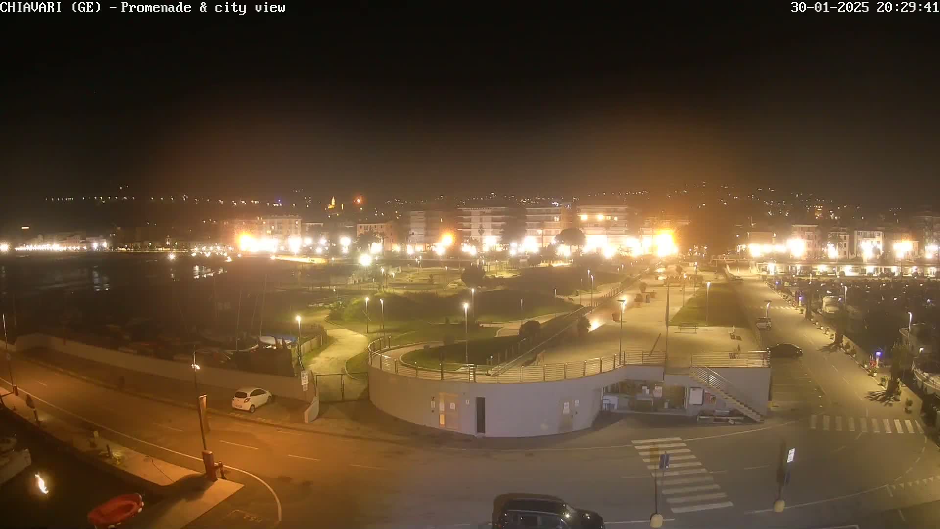 A nighttime view of a city's waterfront promenade and marina, brightly lit, with a calm body of water visible in the distance.