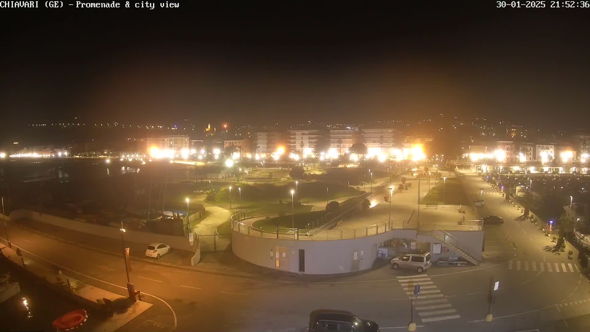 A nighttime aerial view of a waterfront promenade and city, illuminated by streetlights, with cars parked in a lot.