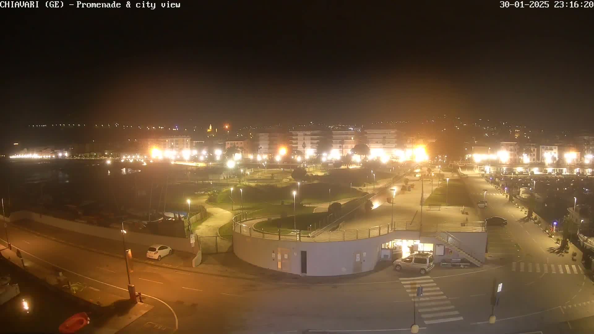 A nighttime, illuminated view of a harbor promenade and city, with a few cars parked and some boats visible in the marina.