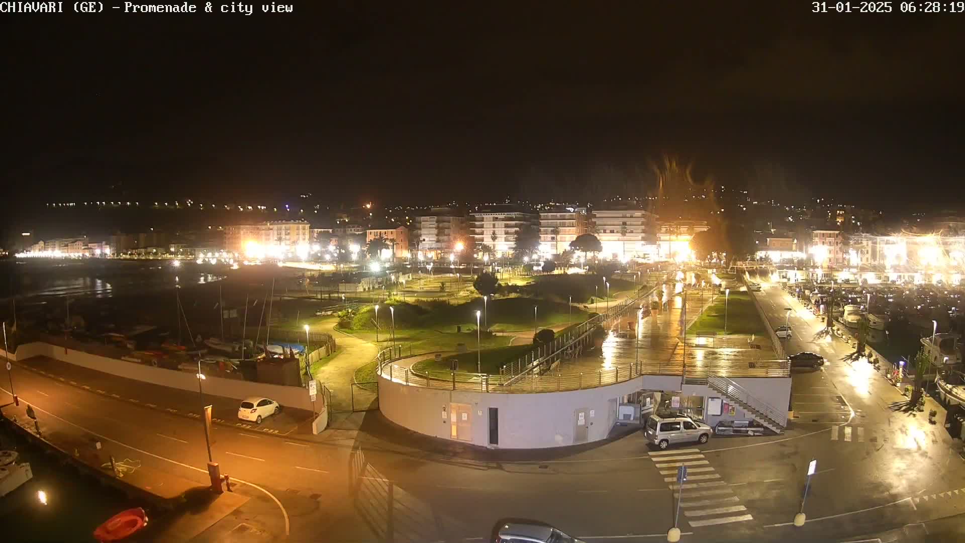 A nighttime view of a waterfront promenade and city, illuminated by streetlights, with wet pavement indicating recent rain.
