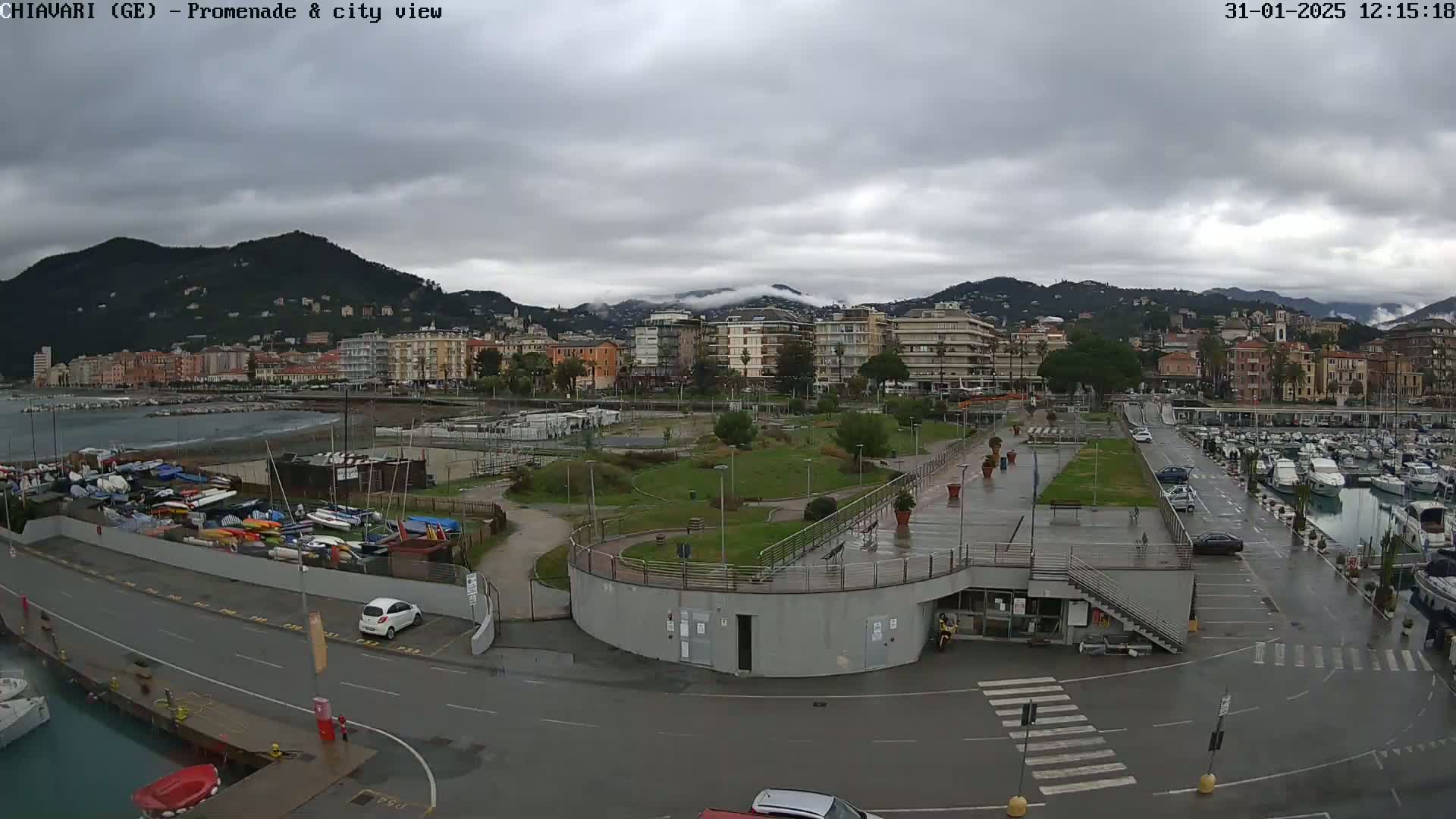 A wet waterfront promenade and marina, with a city and mountains in the background under a cloudy sky.