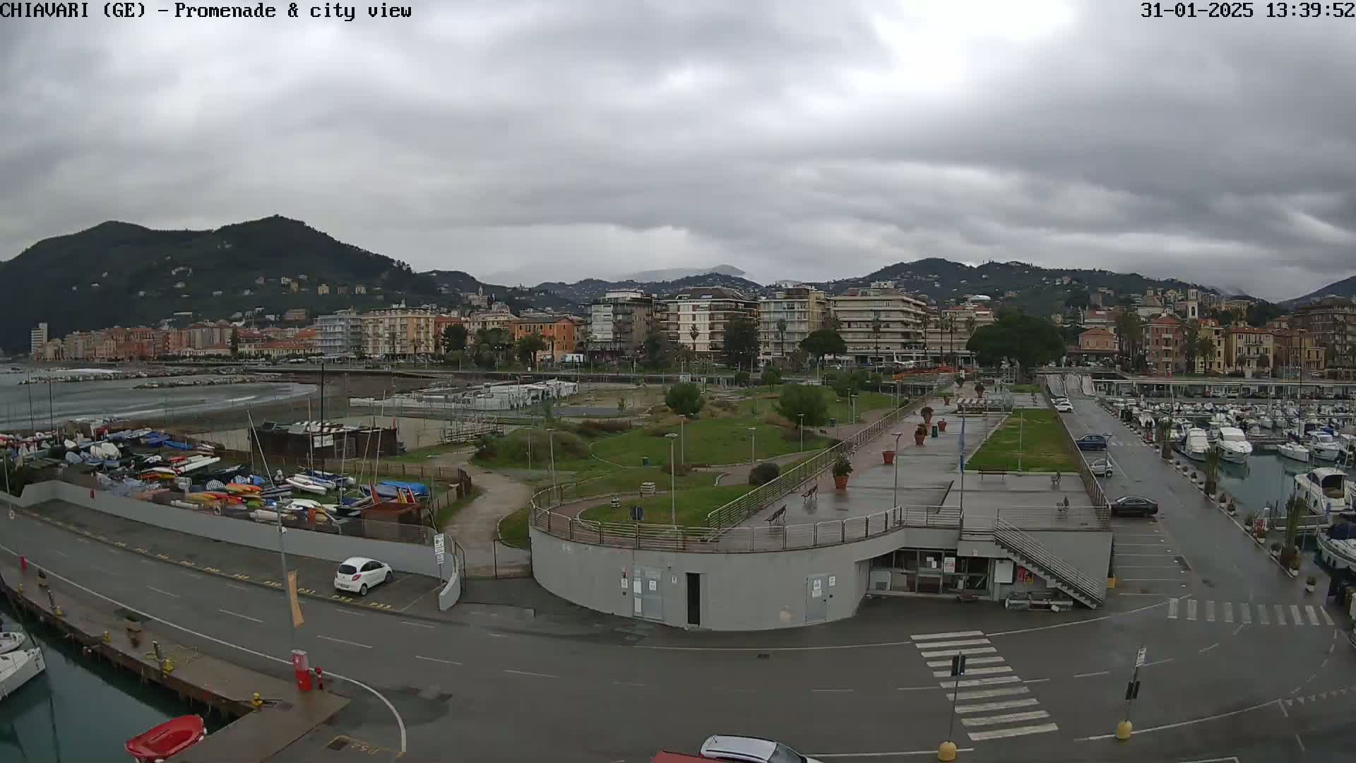 A coastal city scene under a cloudy sky shows a harbor filled with boats, a grassy park, and buildings along a waterfront promenade.