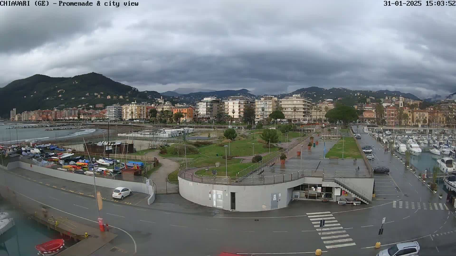 A wet coastal town is viewed from above, showing a marina, promenade, park, and buildings nestled against mountains under a cloudy sky.