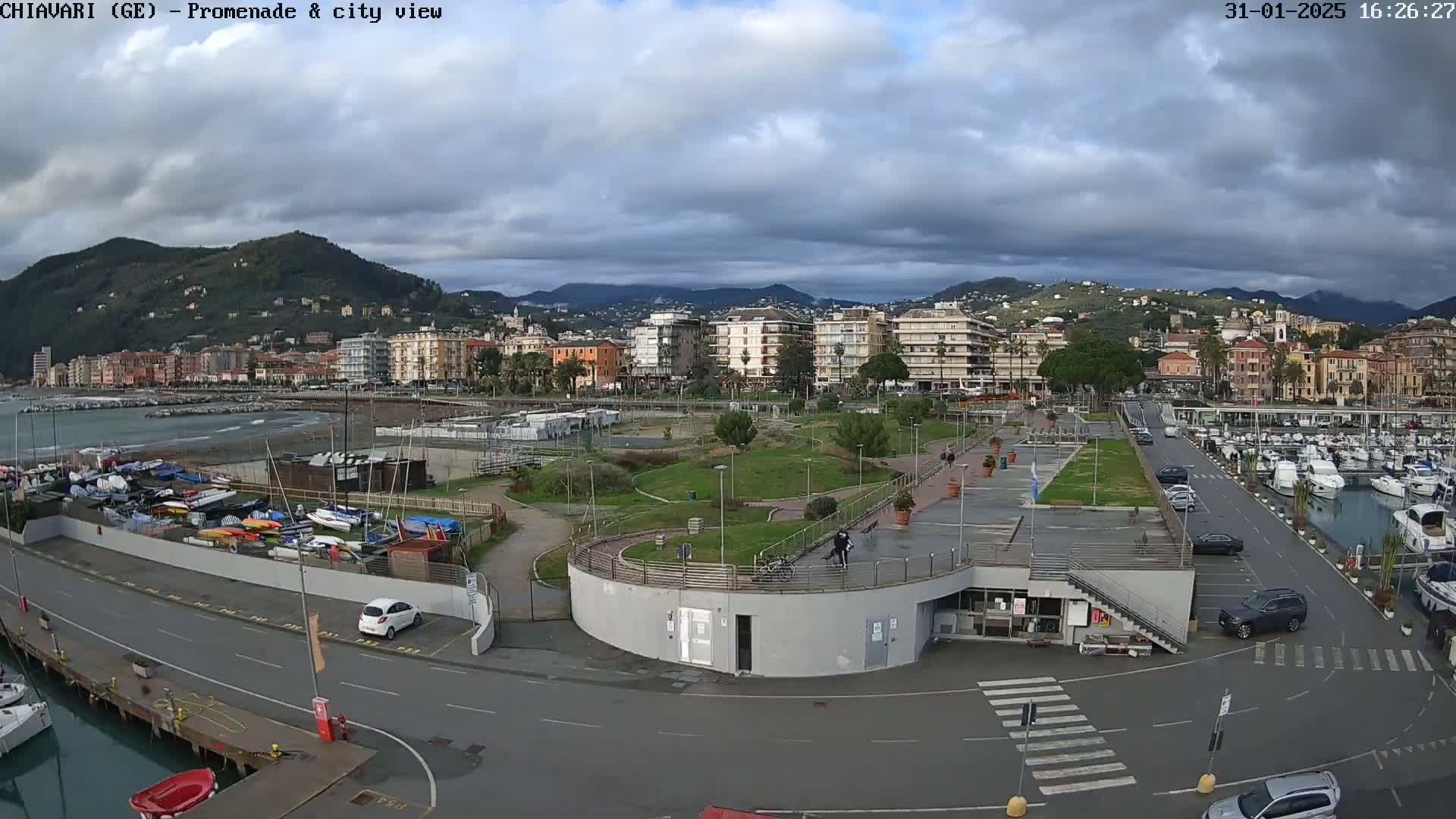 An aerial view of a coastal city on an overcast day shows a marina, promenade, park, and numerous buildings stretching into the distance against a backdrop of mountains.