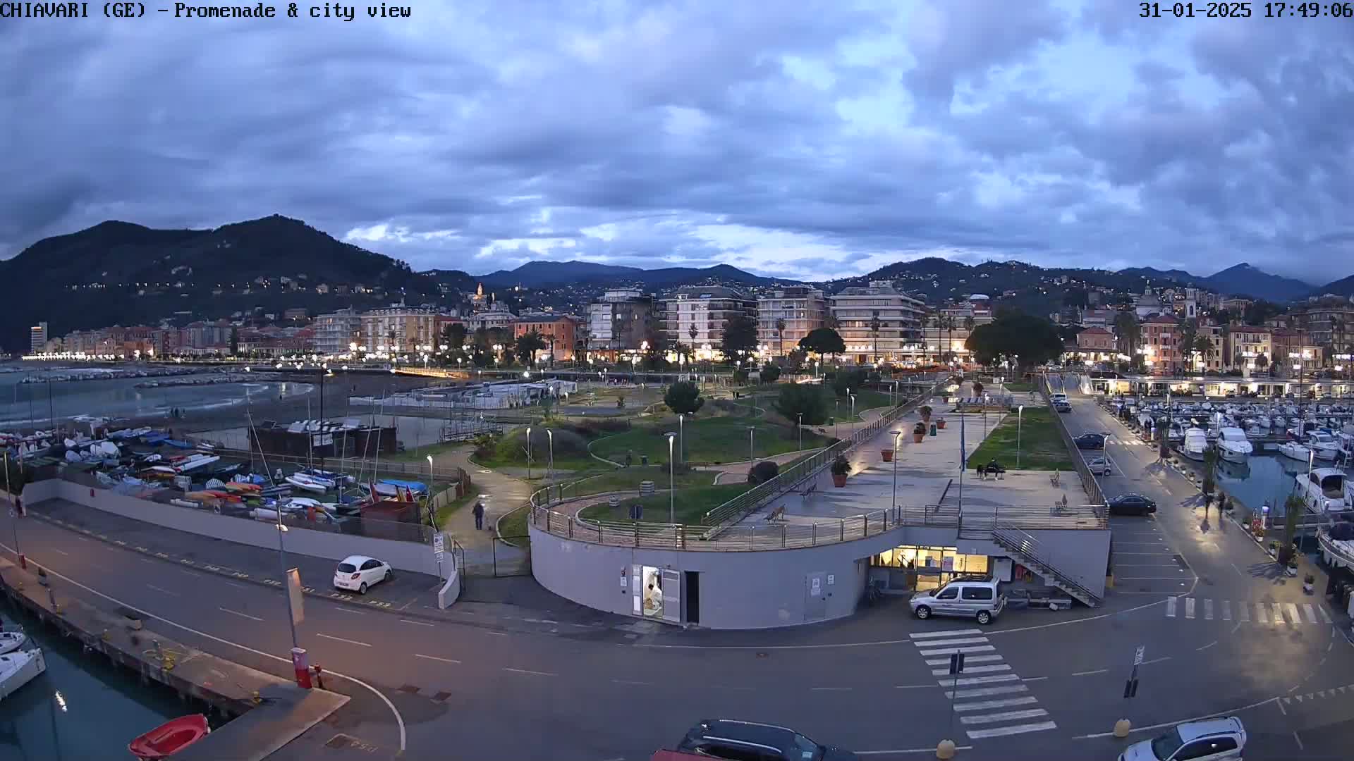 A coastal city at dusk under a cloudy sky, features a marina filled with boats, a promenade with a park, and buildings extending into the background with mountains visible beyond.