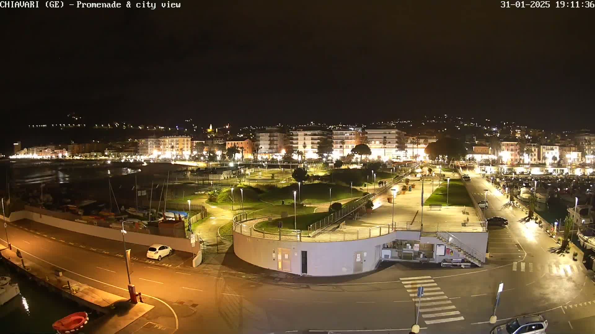 A nighttime aerial view of a coastal city's promenade and harbor, illuminated by streetlights, showing a park-like area and a marina with boats.
