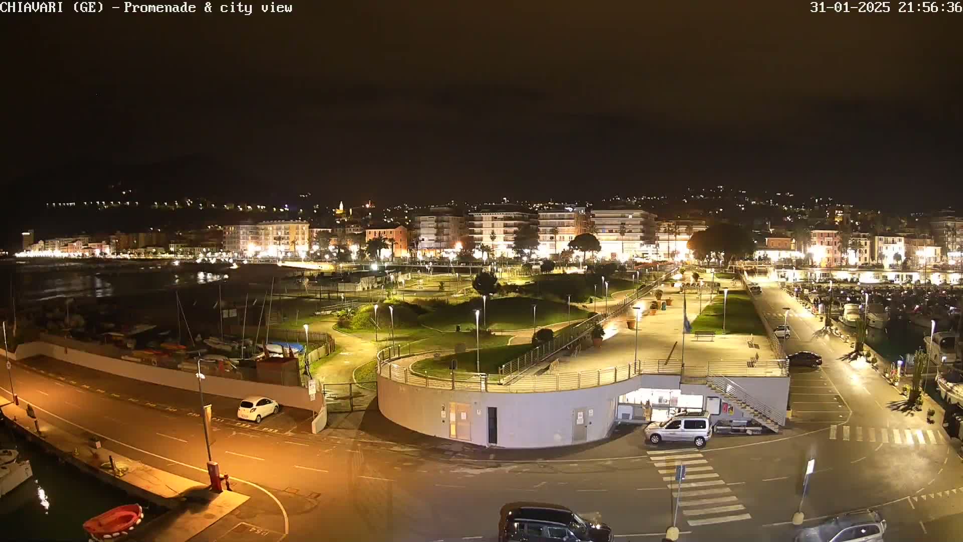 A nighttime view of a coastal city's illuminated promenade and harbor, featuring a park, parked cars, and boats.