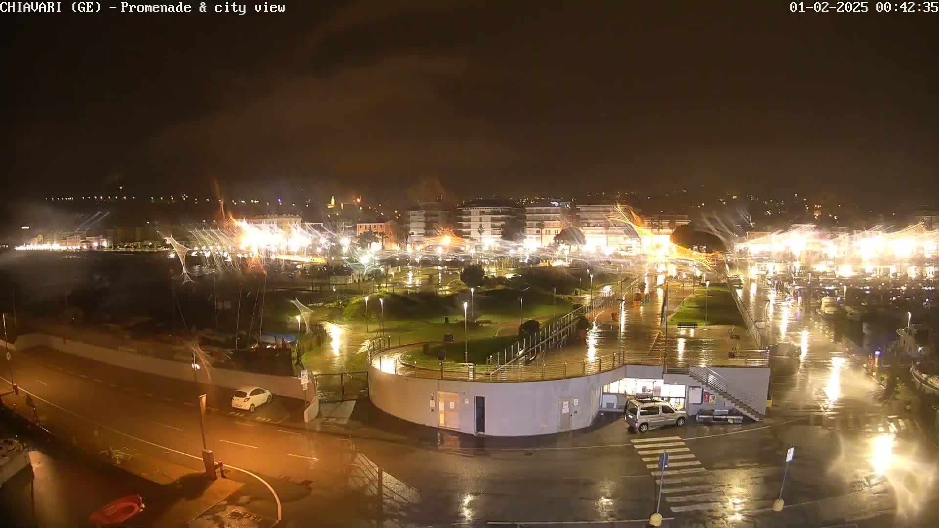A nighttime view of a rain-soaked city promenade and harbor, illuminated by lights, with water features creating streaks of light across the scene.