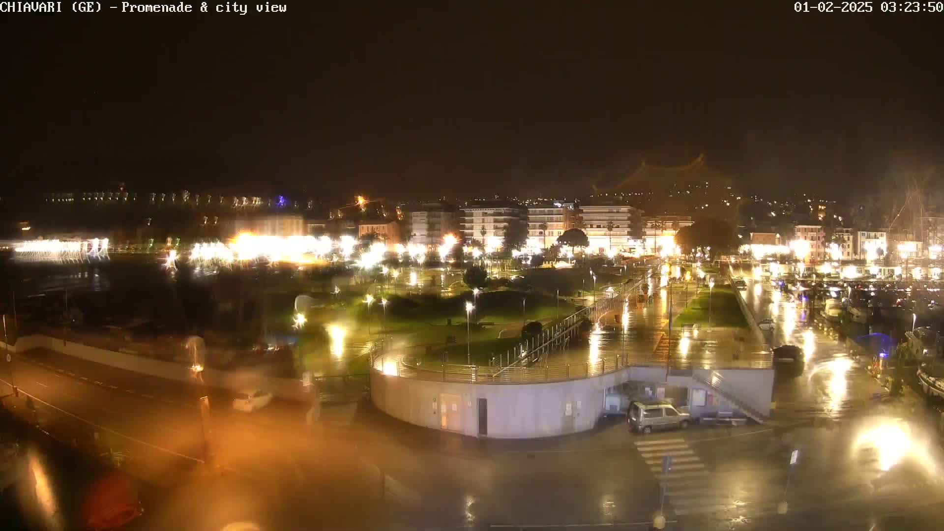 A nighttime, rain-soaked city promenade and waterfront area, illuminated by streetlights, shows a few cars and buildings in the background.