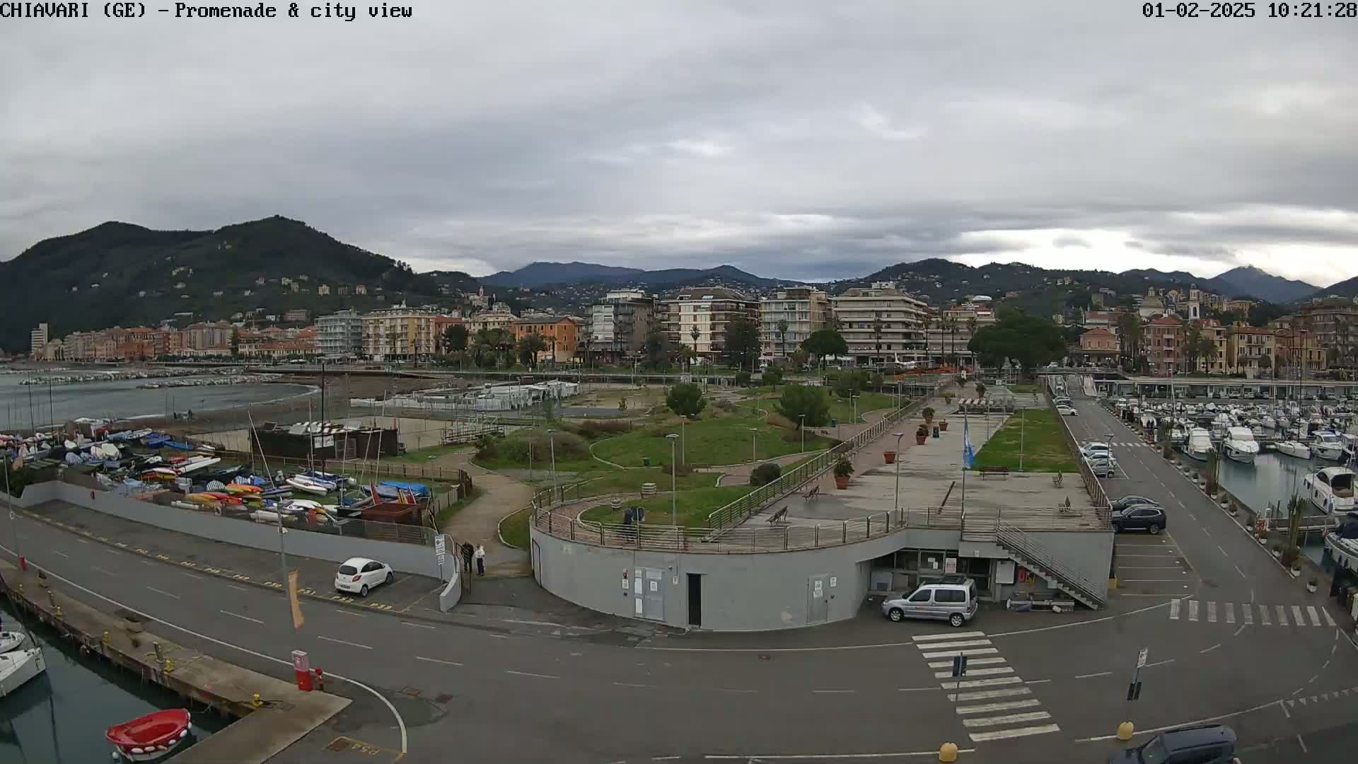 A coastal city with a harbor filled with boats, a promenade with green spaces, and buildings on a hillside under a cloudy sky.