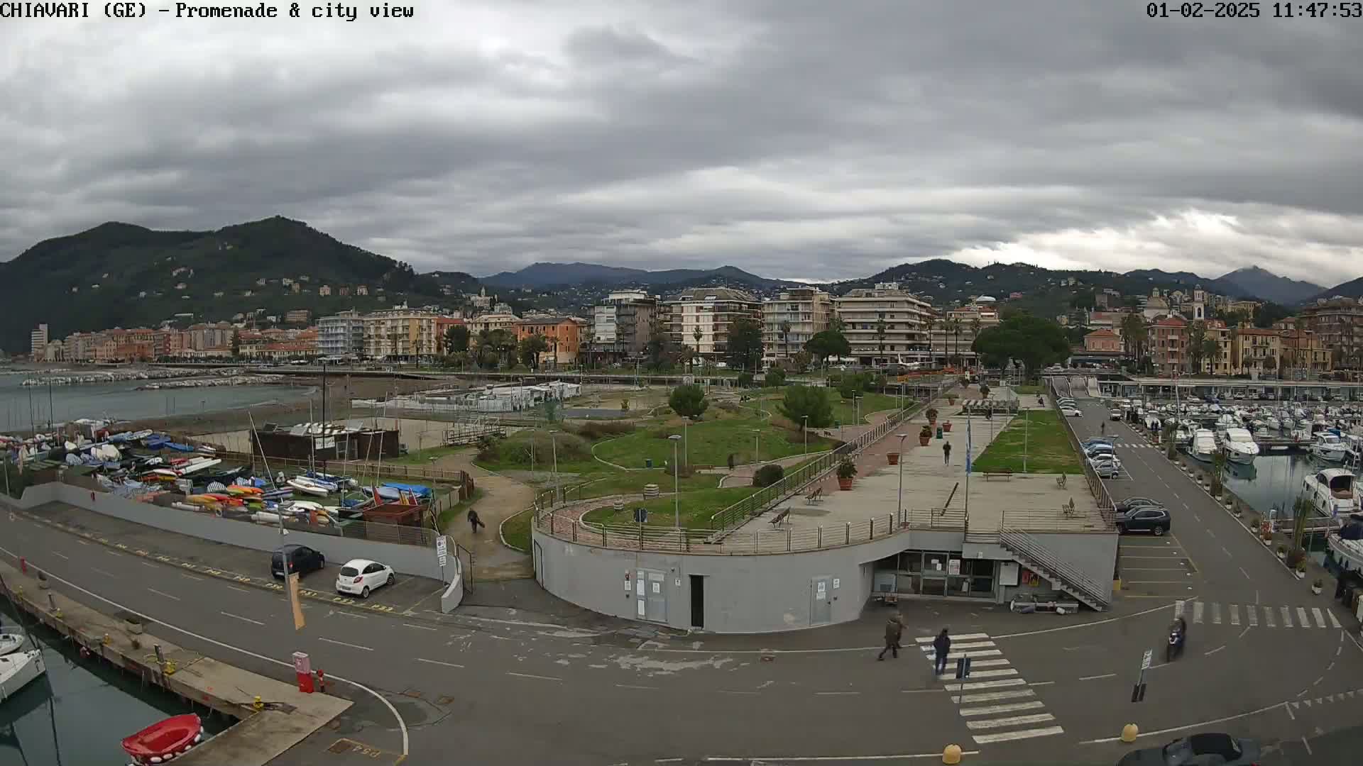 A coastal city scene under a cloudy sky shows a harbor filled with boats, a promenade with green landscaping, and buildings rising behind it on a hillside.