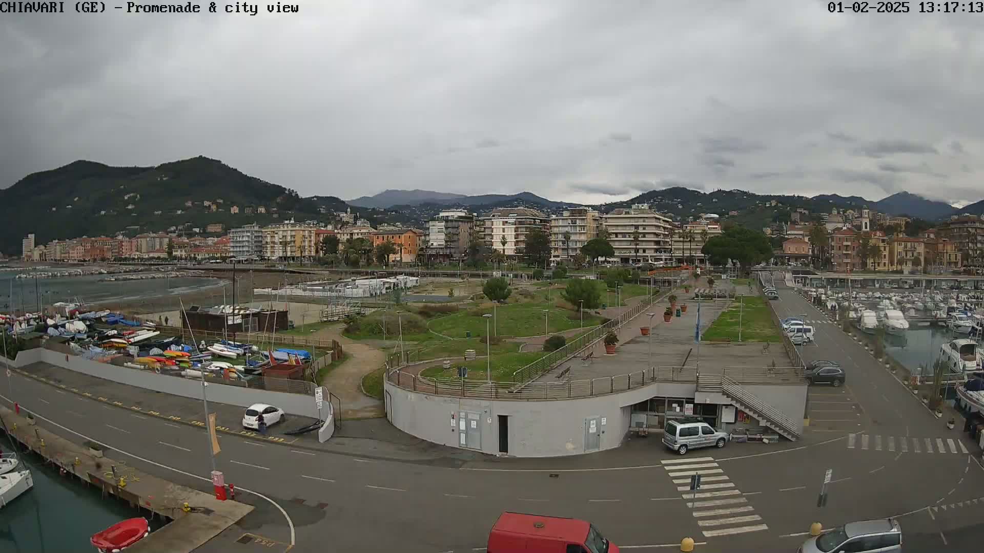 A seaside town with a harbor full of boats, a promenade, and green spaces sits under a cloudy sky.