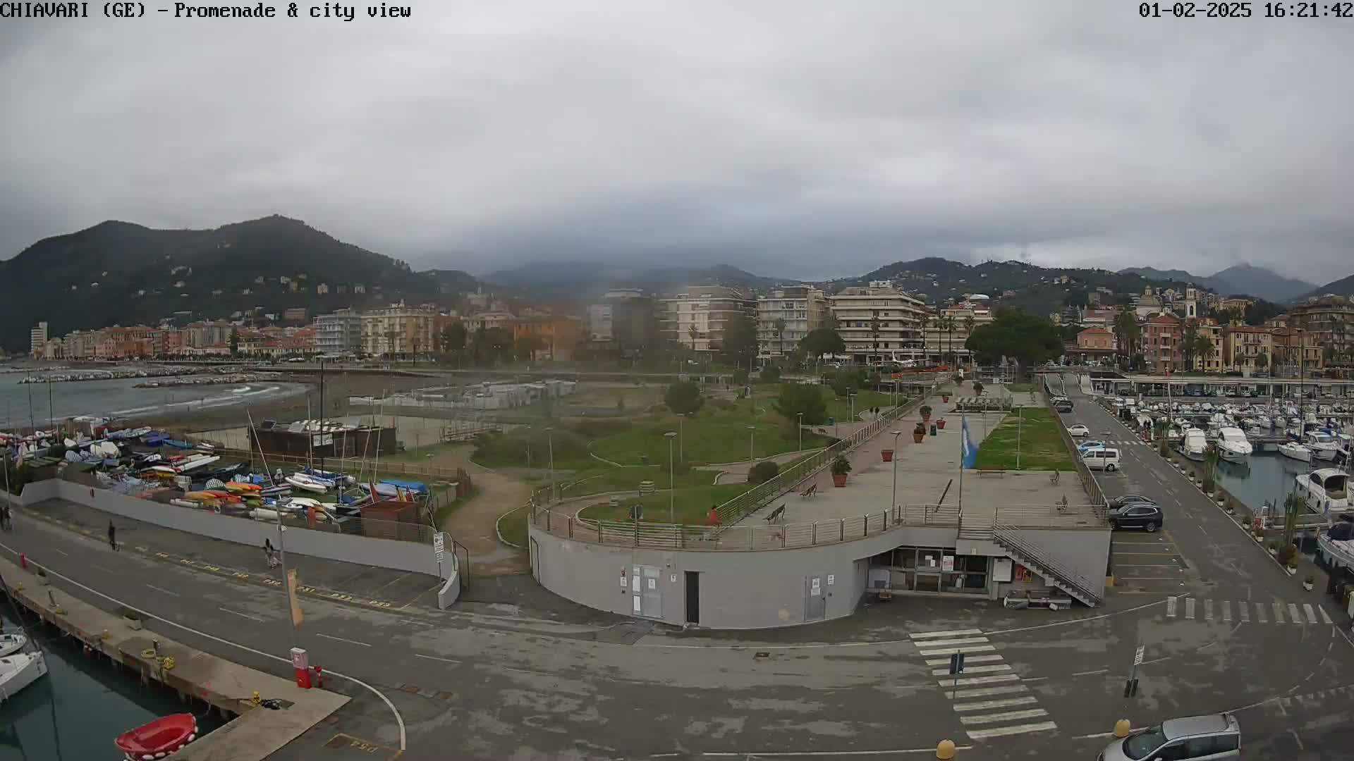 An overcast day reveals a coastal city with a marina, promenade, and park, all situated against a backdrop of mountains.