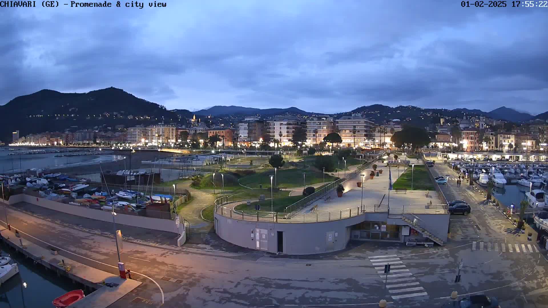 A coastal city at twilight, with a marina filled with boats, a promenade, and a park, all under a partly cloudy sky.