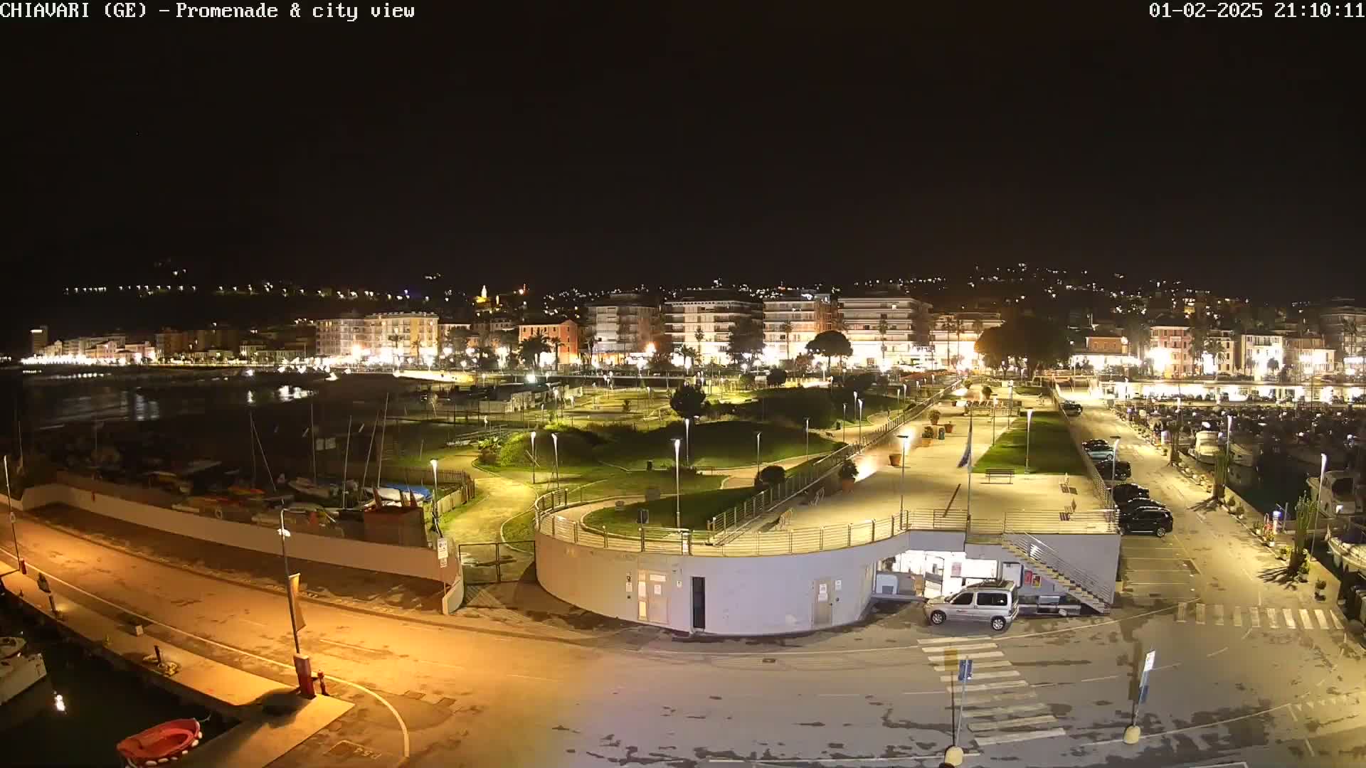 A nighttime, high-angle view of a waterfront promenade and city, illuminated by streetlights, showing a calm body of water, boats docked, and a park-like area with winding pathways.
