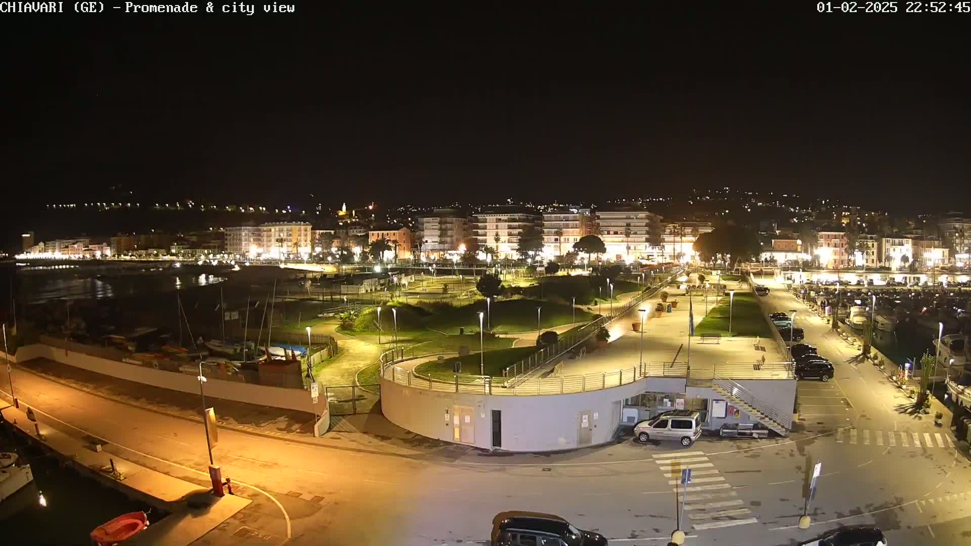 A nighttime view of a city's illuminated waterfront promenade, harbor, and parking lot, with boats moored and cars parked.
