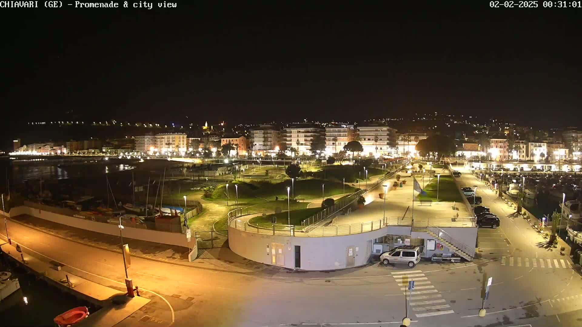 A nighttime view of a city's waterfront promenade and harbor, illuminated by artificial lights, with parked cars and boats visible.