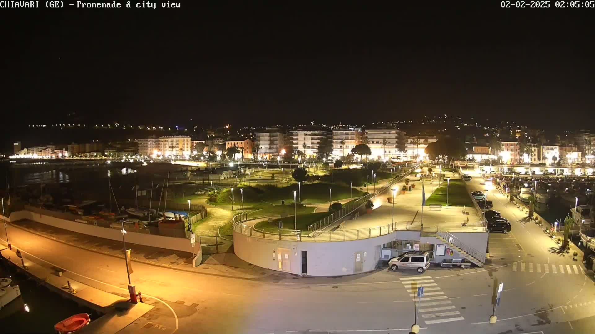 A nighttime, aerial view of a city's illuminated waterfront promenade, featuring a marina with boats, a park, and a parking lot.