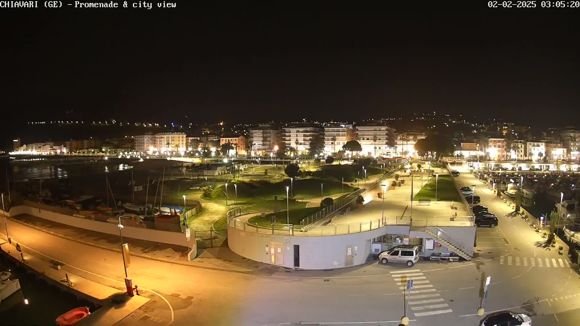 A nighttime, illuminated view of a waterfront promenade and city, with a harbor containing boats and a parking lot.