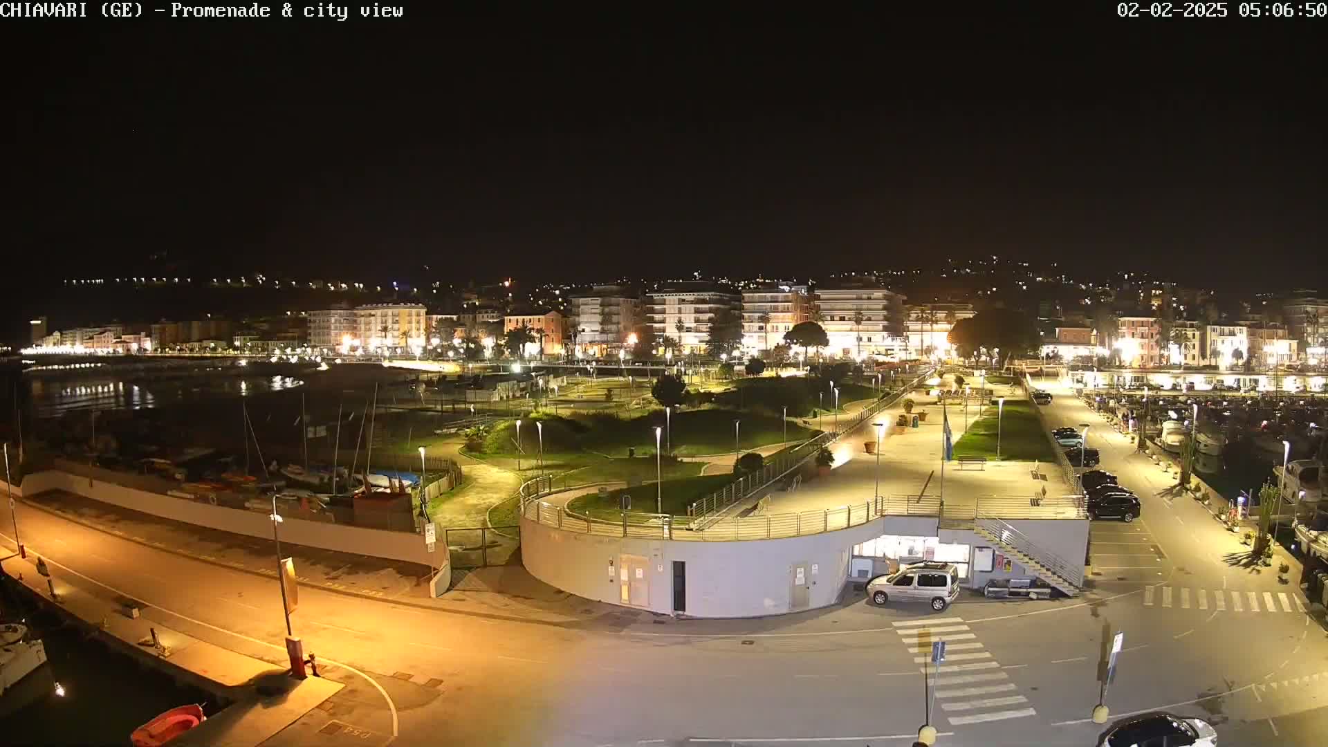 A nighttime, brightly lit view of a harbor promenade and city, with boats moored in the water and cars parked in a lot.