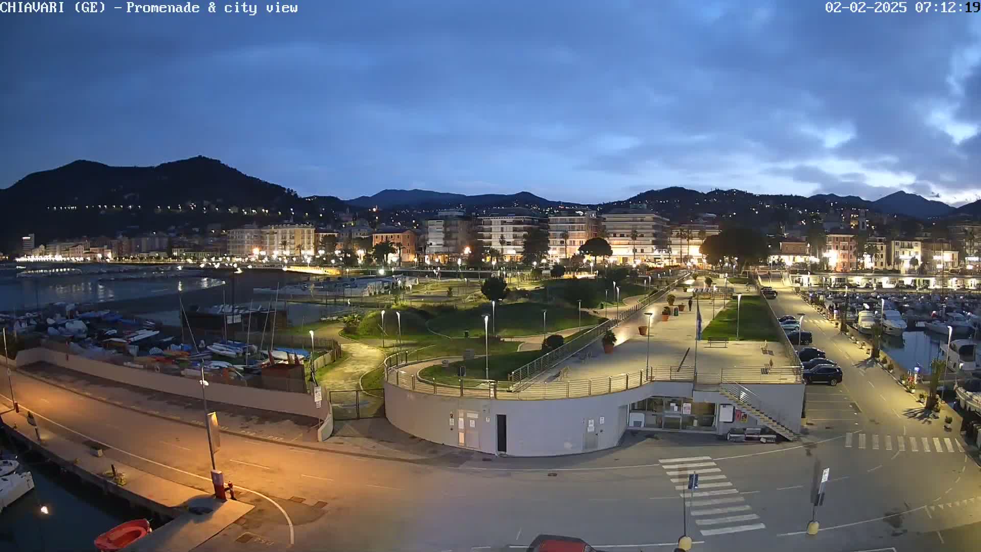 An illuminated waterfront promenade and city at dusk, under a mostly clear sky, showing a harbor with boats, a park, and buildings stretching into the background hills.