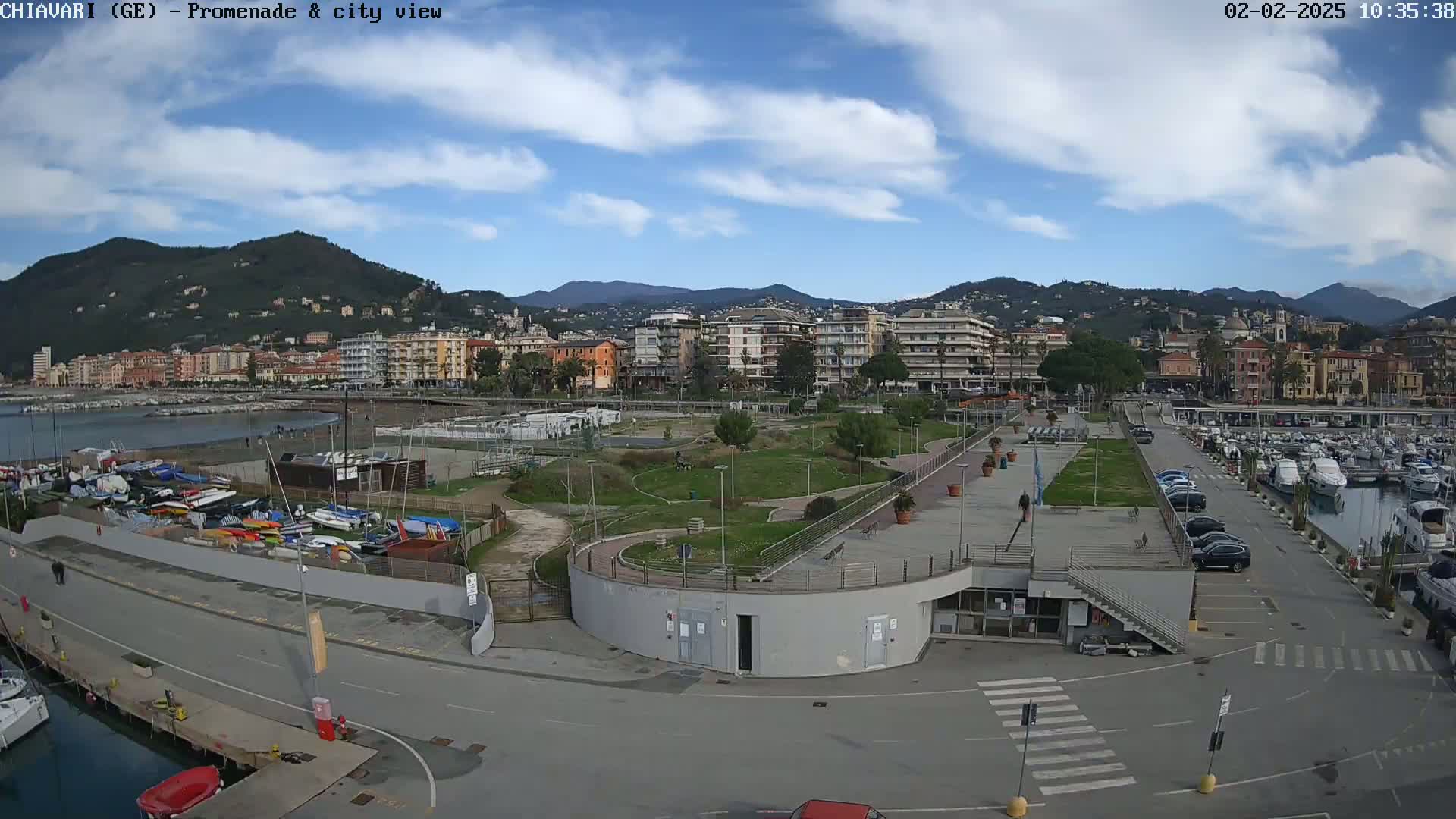 A coastal city scene under a partly cloudy sky shows a marina with numerous boats, a paved promenade, and a park-like area, all situated before a backdrop of buildings and hills.