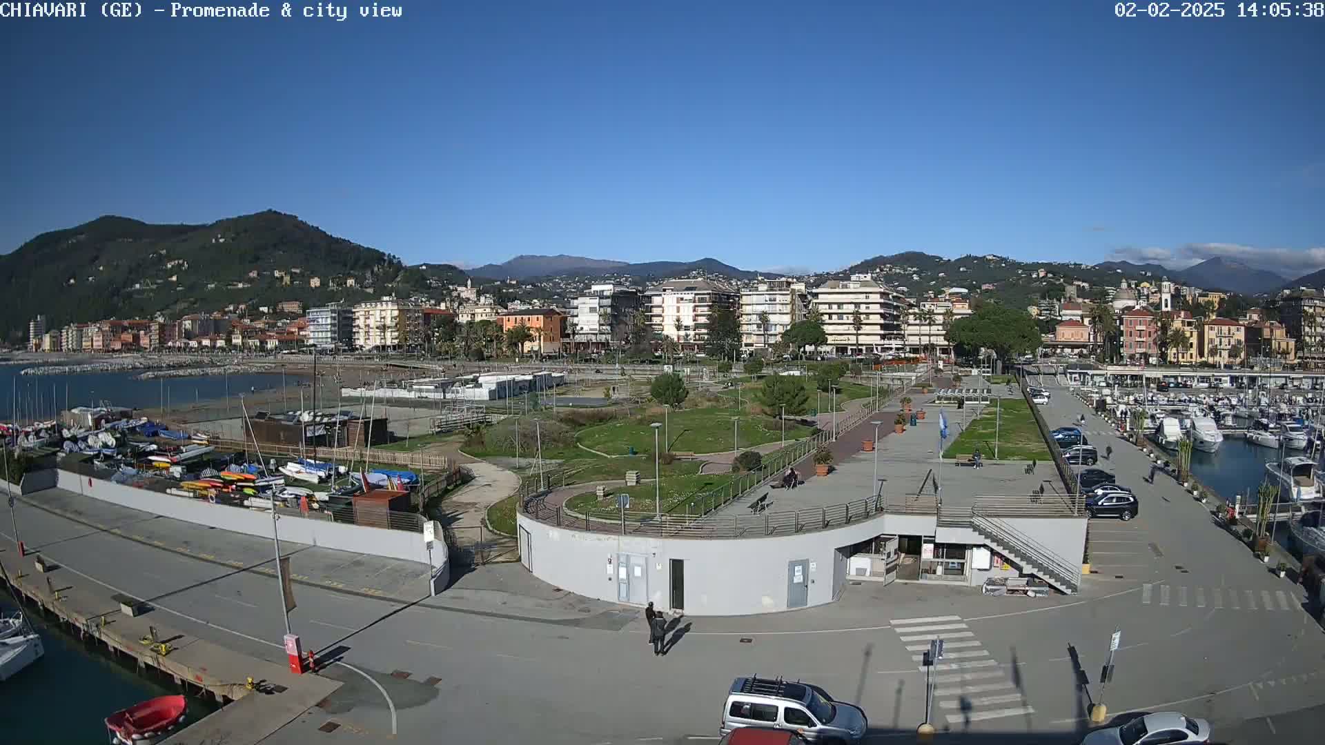 An aerial view of a waterfront promenade with a marina, park, and cityscape in the background under clear blue skies.