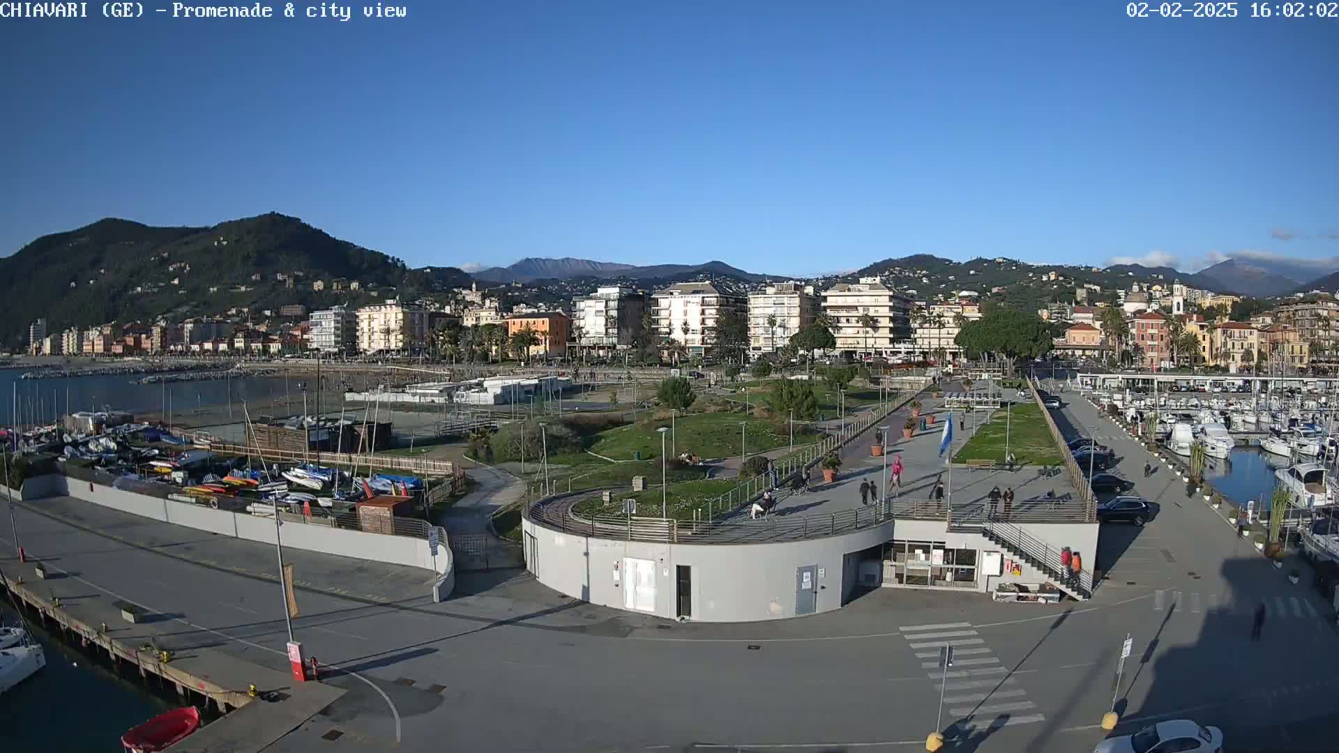 A waterfront promenade with a marina and a city in the background under a clear blue sky.