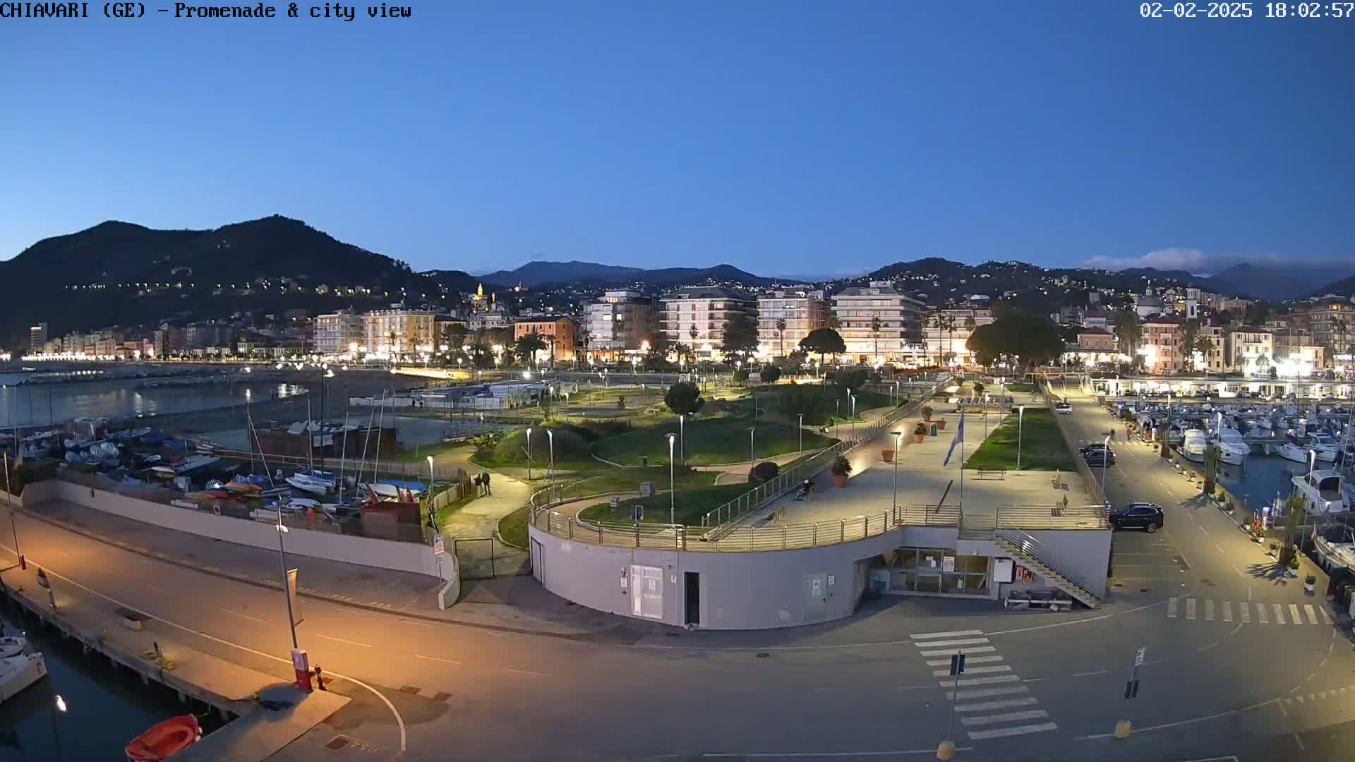 A coastal city at twilight, with a marina filled with boats, a park, and buildings illuminated against a backdrop of mountains.