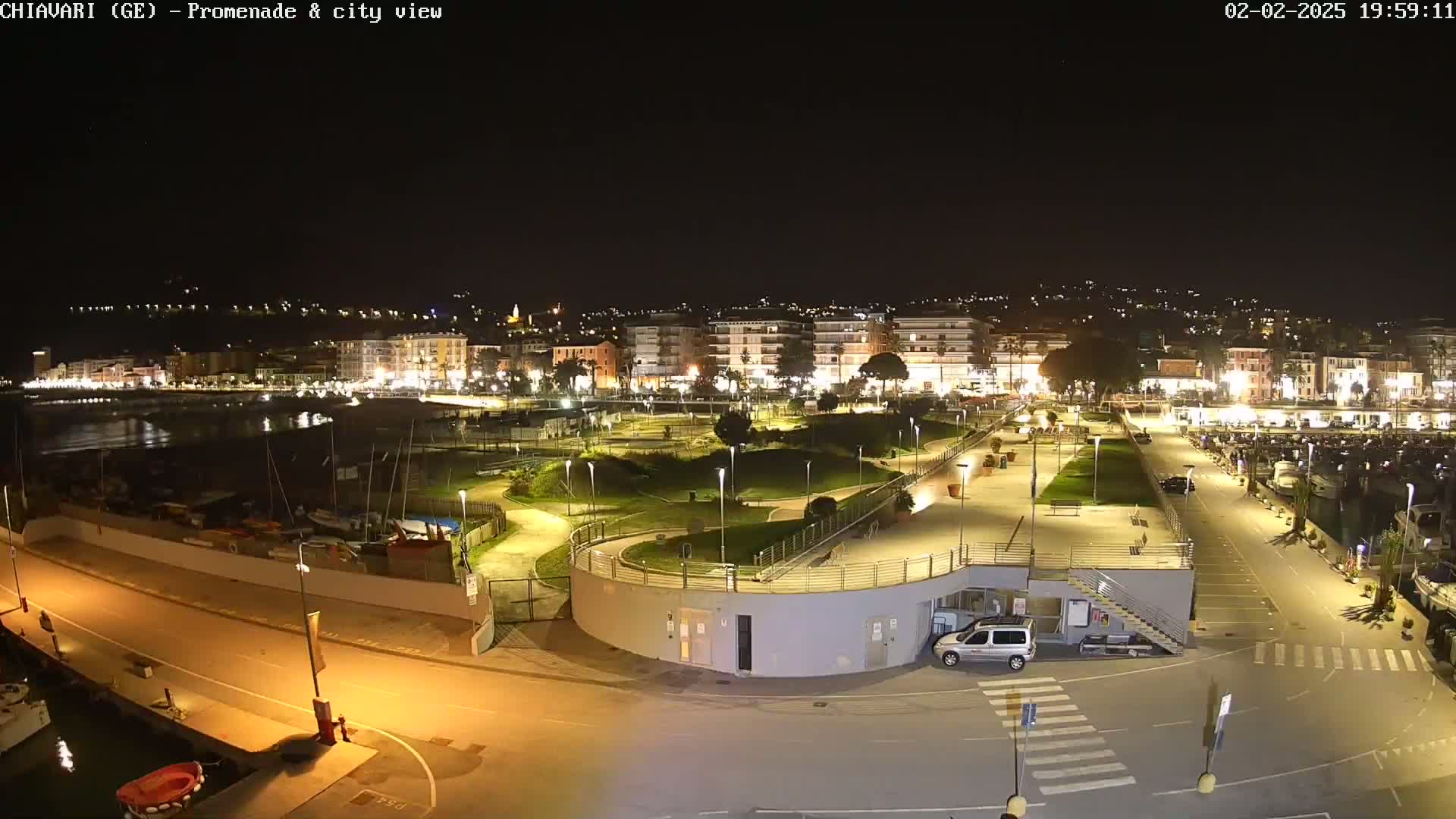A nighttime, aerial view of a city's waterfront promenade and harbor, featuring lit buildings, a park, and boats moored at the docks.