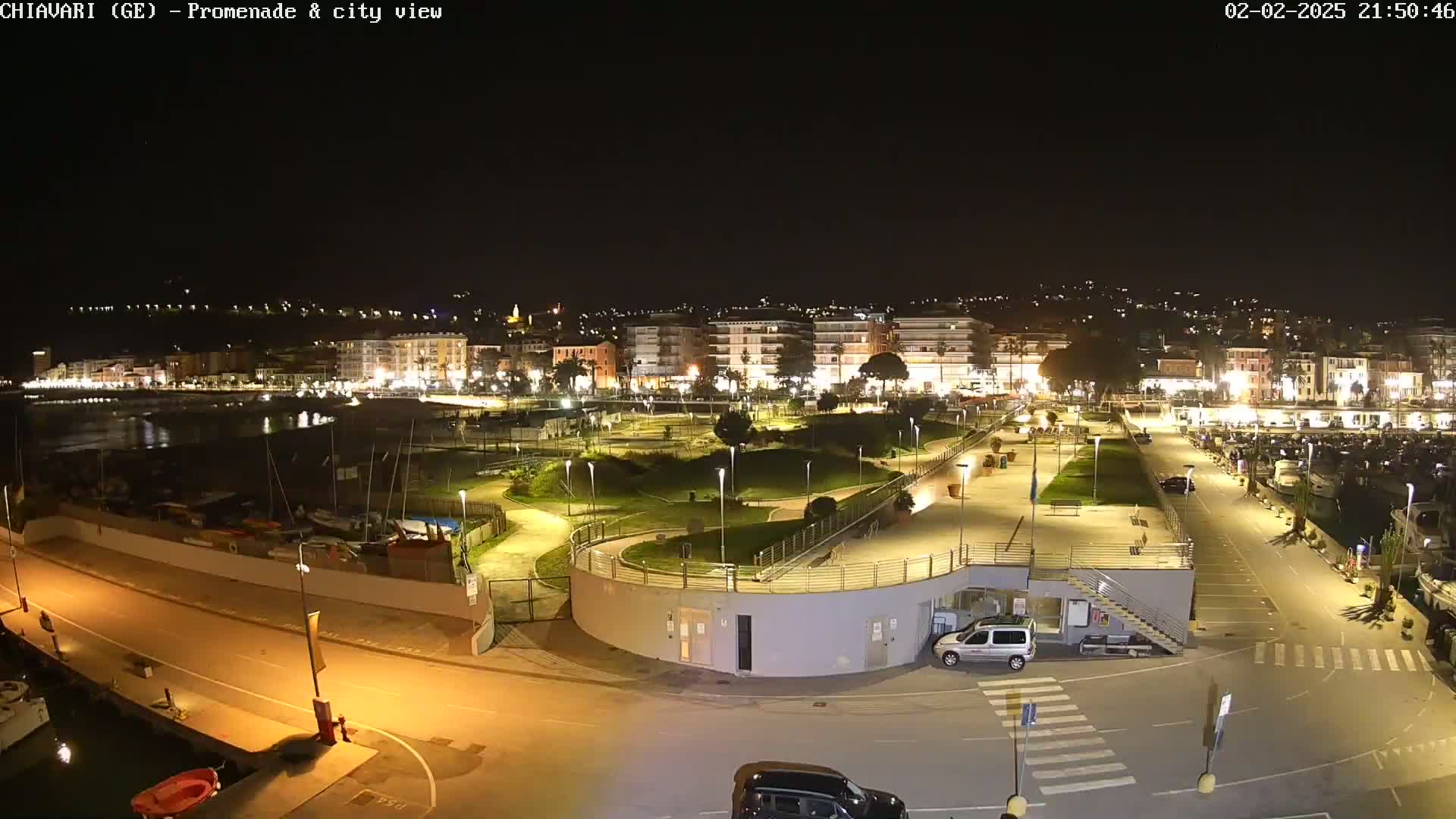 A nighttime view of a brightly lit waterfront promenade and city, with a marina and parked cars visible.