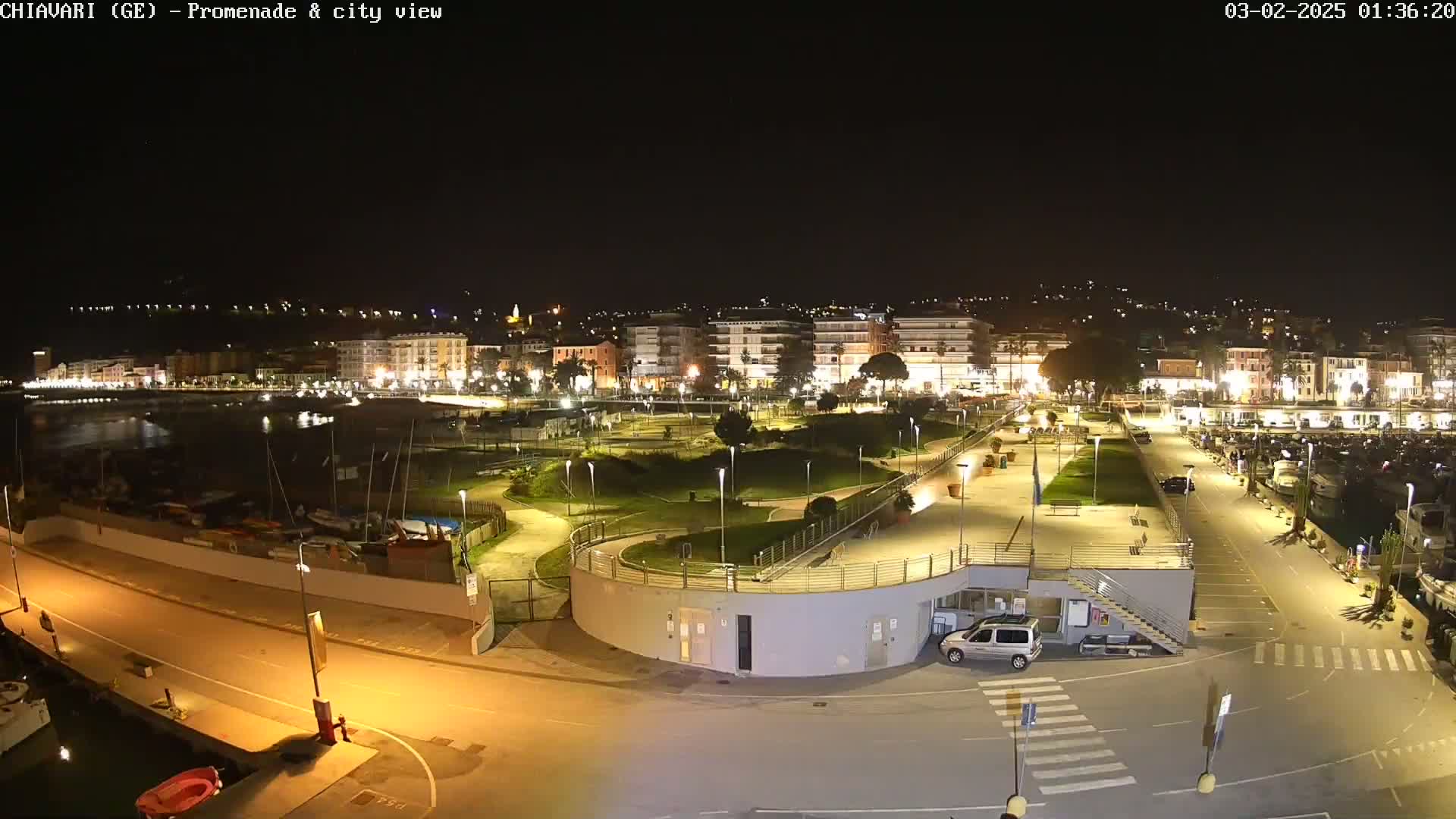 A nighttime aerial view of a waterfront promenade and city, illuminated by streetlights, with a marina filled with boats.