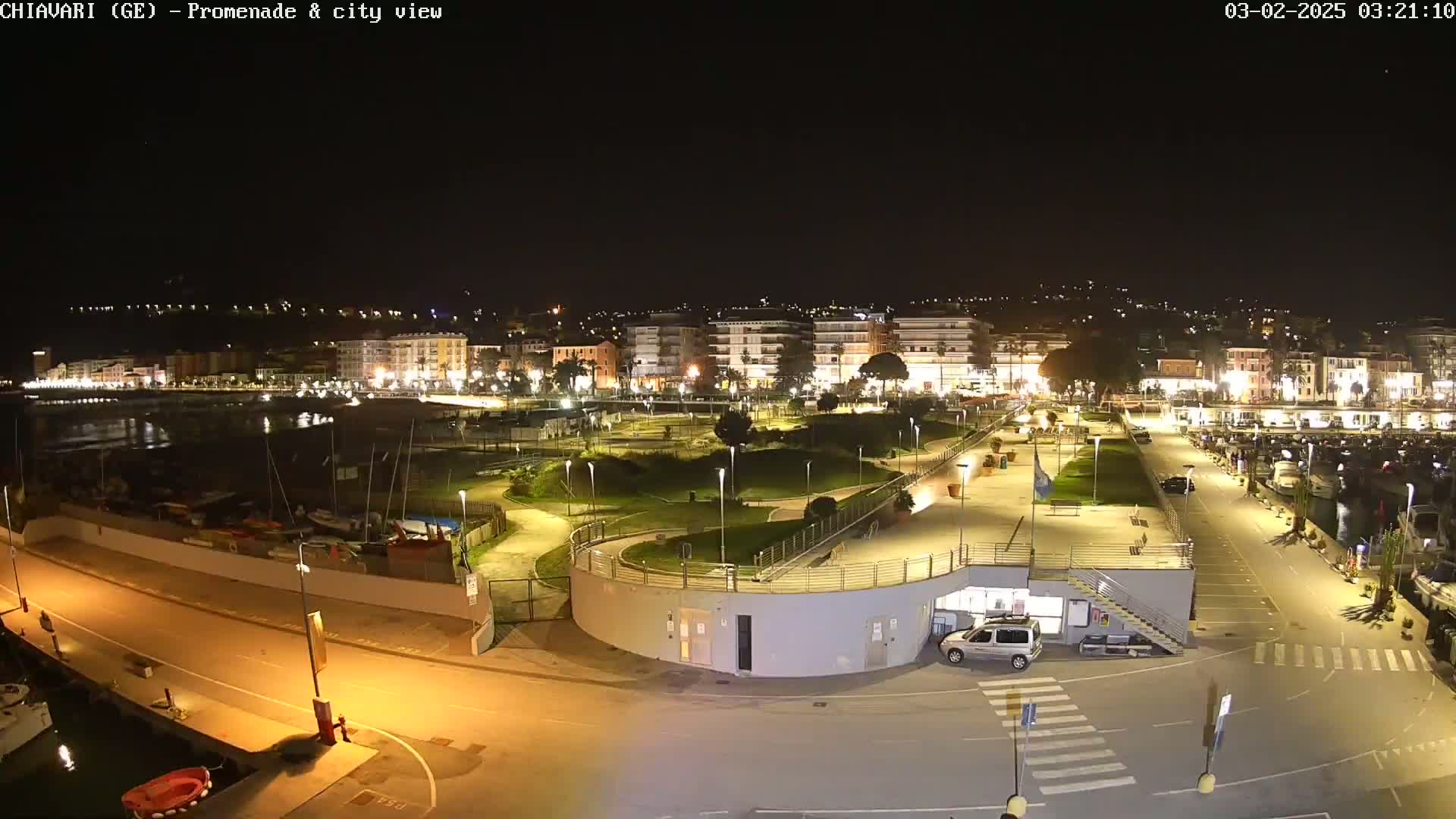 A nighttime aerial view of a city's waterfront promenade and harbor, featuring lit buildings, a park, and boats moored along the water's edge.