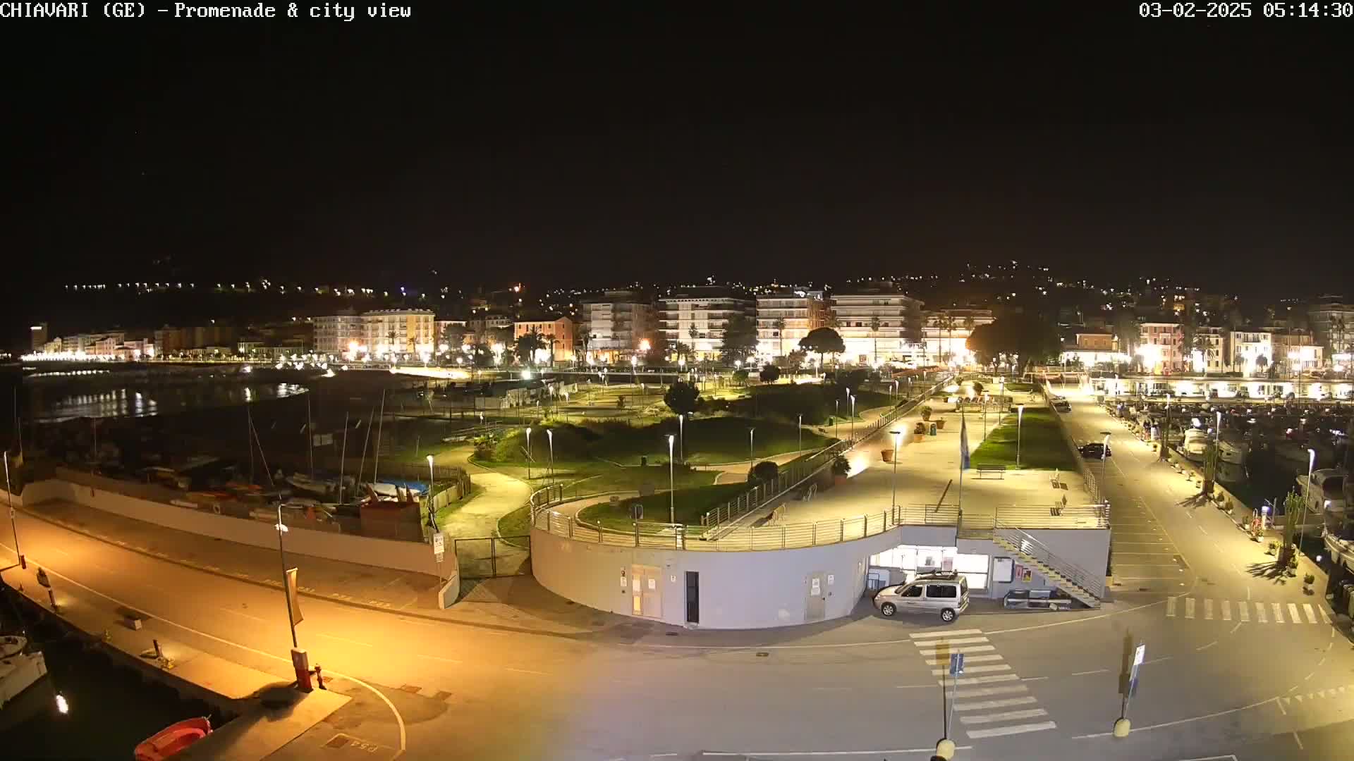 A nighttime view of a city's illuminated waterfront promenade and harbor, featuring a park-like area, buildings, and parked cars.