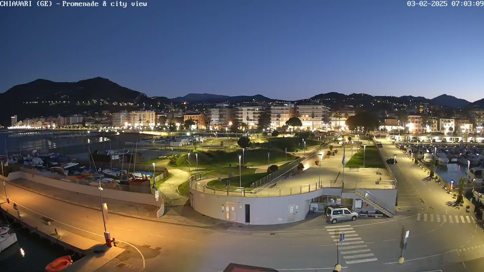 A nighttime view of a coastal city with a marina, promenade, and park, under a clear twilight sky.