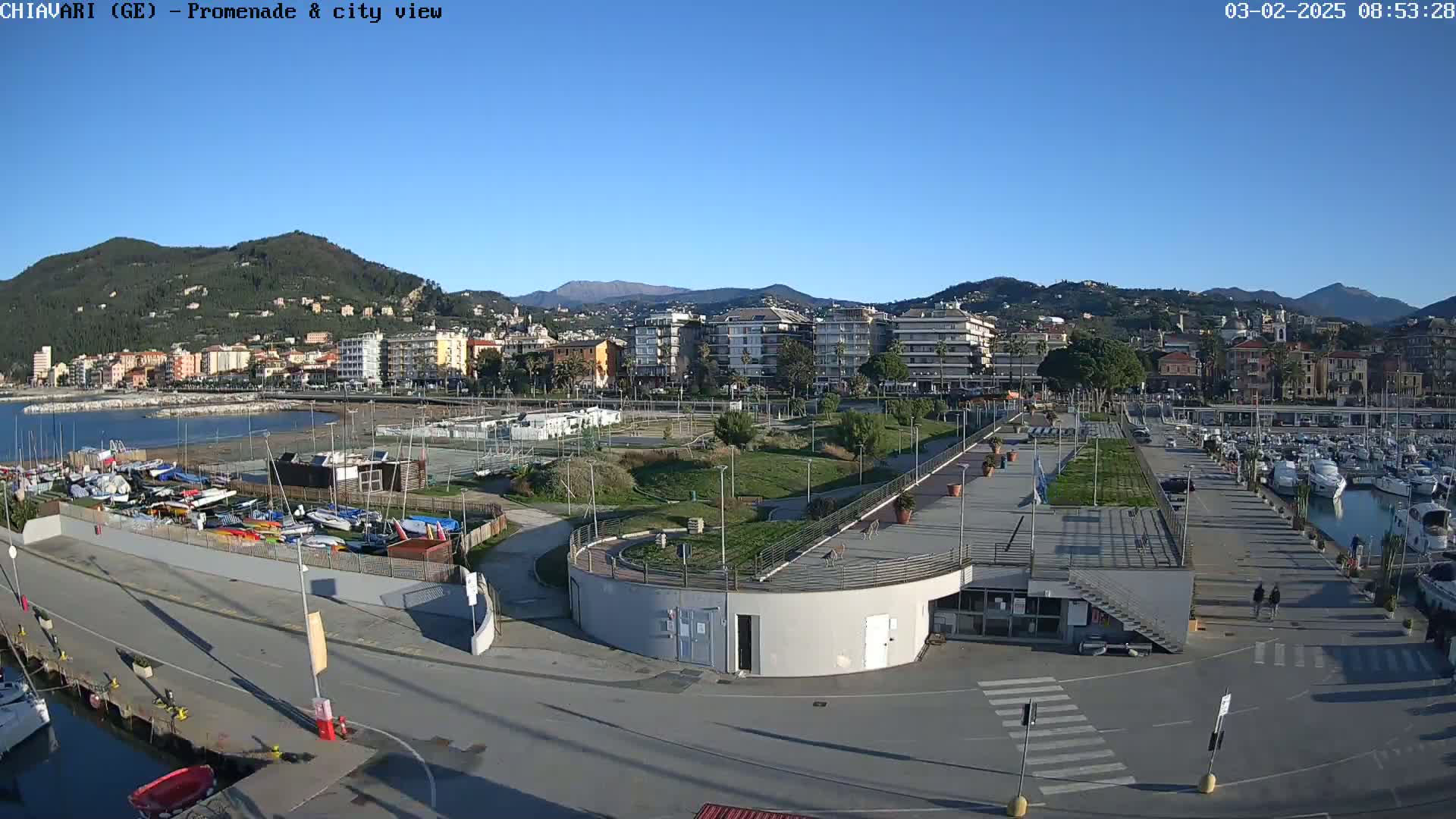 A coastal city scene on a sunny day shows a marina filled with boats, a park-like area with pathways, and buildings extending along the shoreline, backed by hills.