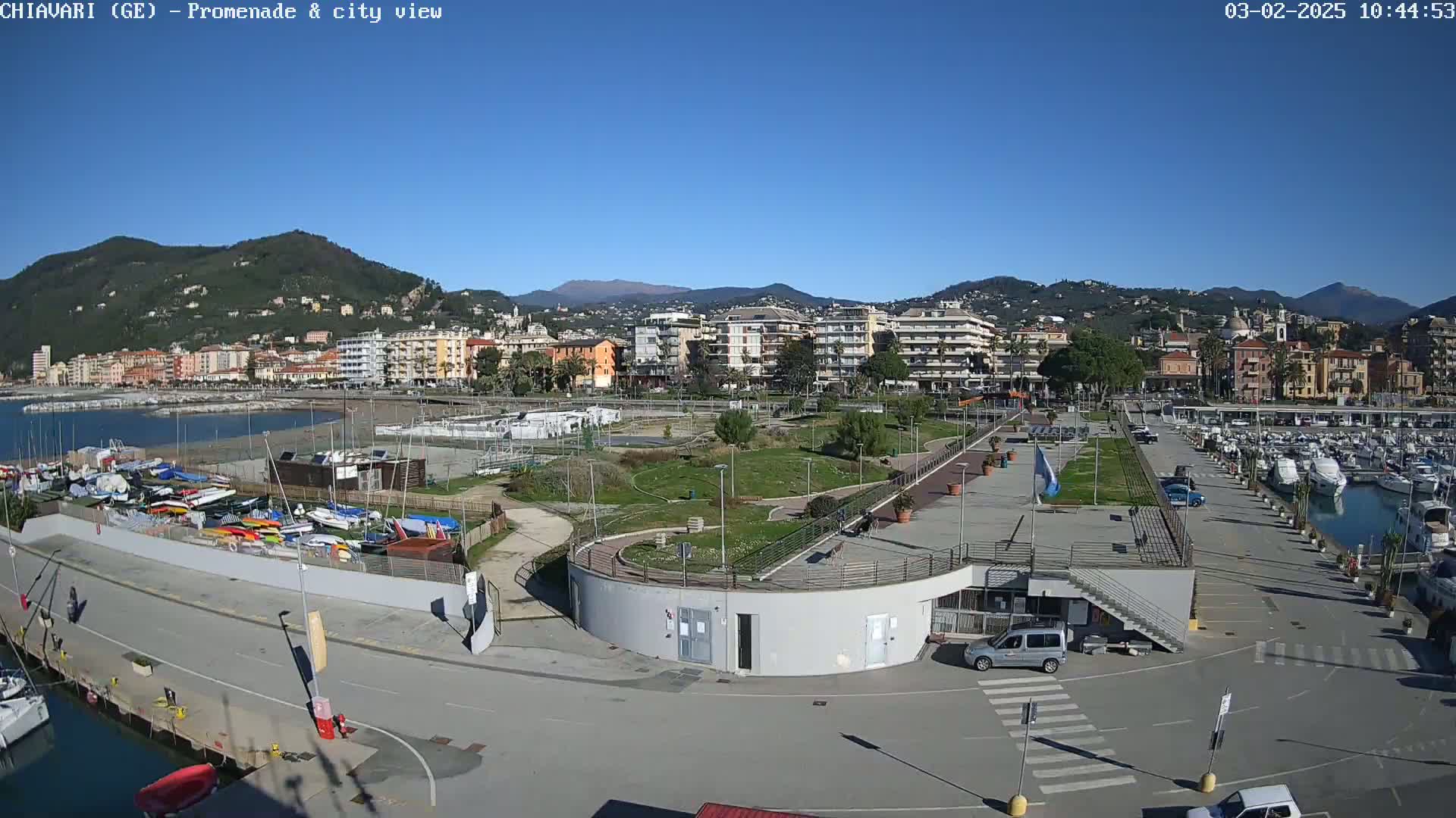 A coastal town with a marina, park, and numerous buildings is seen on a clear, sunny day, backed by mountains.